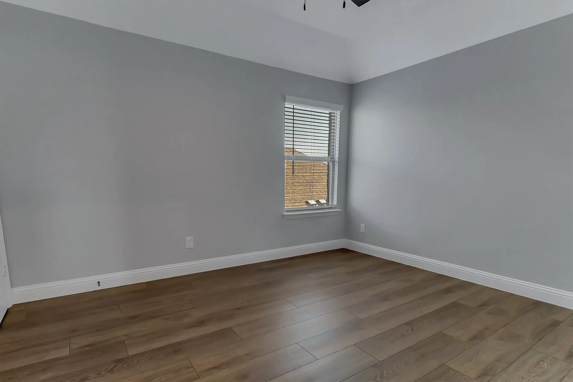 Spacious room featuring wood-finish flooring, light grey walls, white baseboards, and a window with blinds