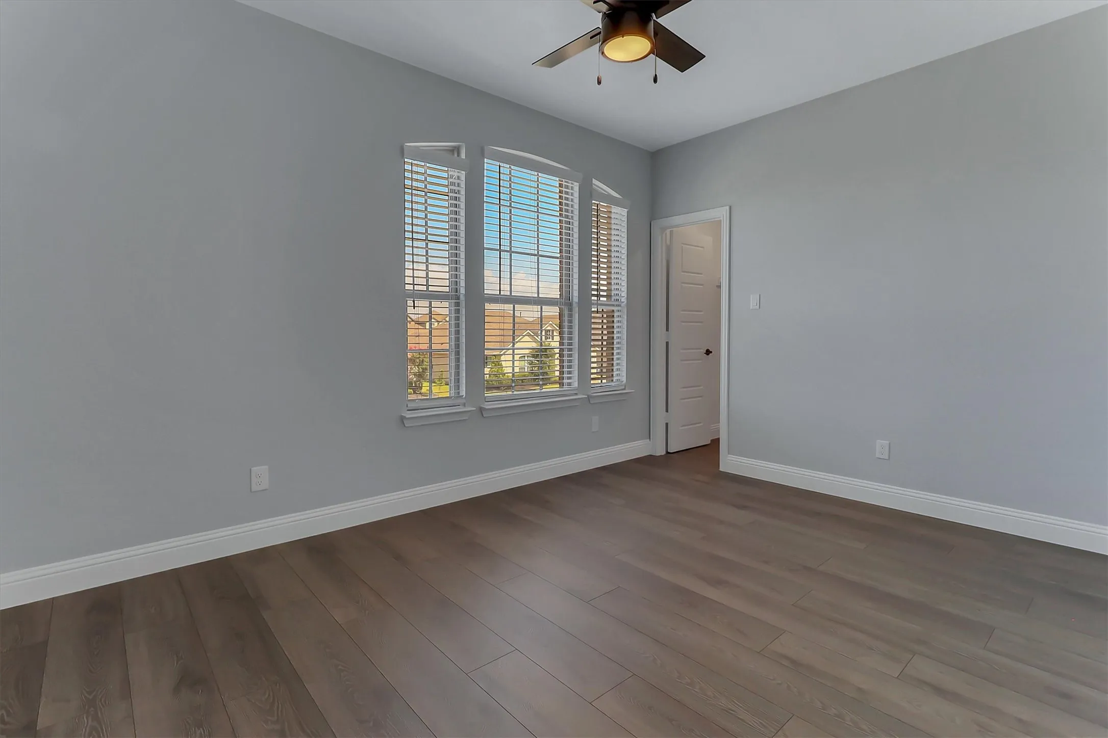 Spacious room featuring wood-finish flooring, light gray walls, a three-panel arched window with blinds, a ceiling fan with integrated lighting, and white baseboards. Walk in closet