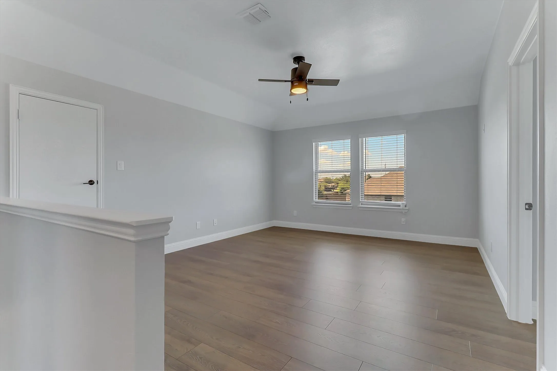 Spacious room featuring wood-finish flooring, two windows with horizontal blinds, a ceiling fan with integrated lighting, a white interior door with black hardware, and crisp white baseboards