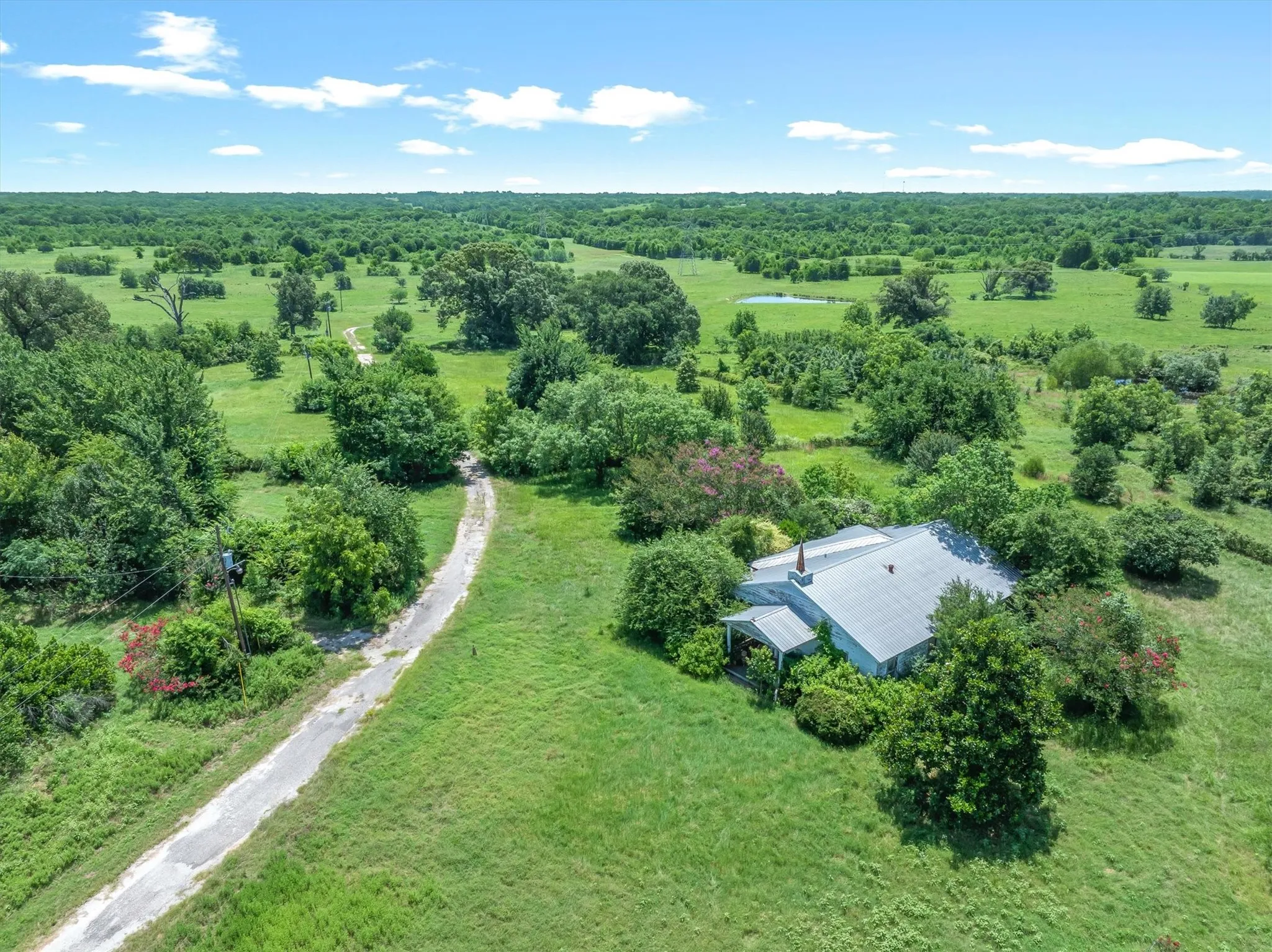 Rural property featuring a single-story residence with a metal roof, surrounded by expansive green fields and mature trees