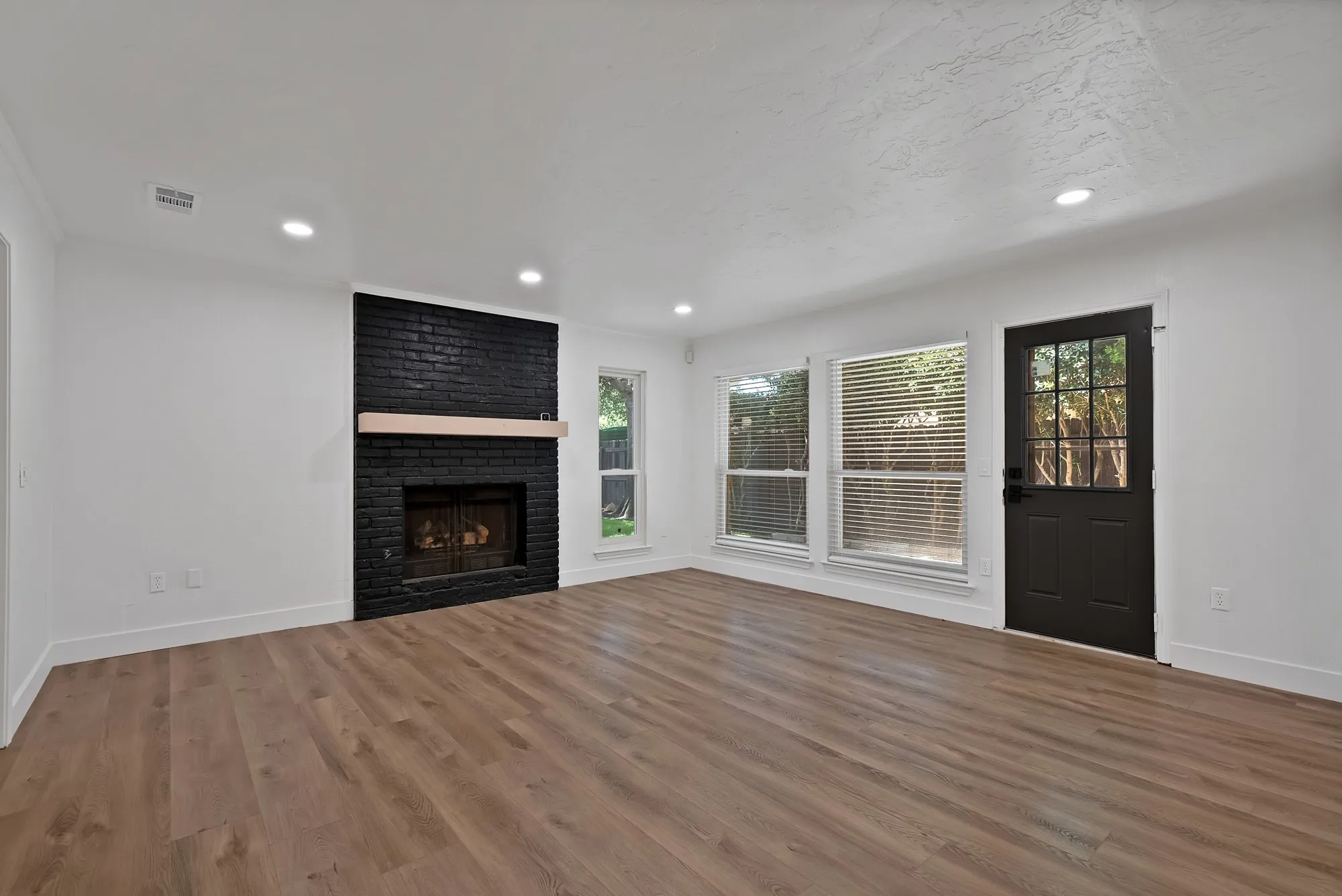 Living space featuring wood-finish flooring, a black brick fireplace with a light wood mantel, recessed lighting, and multiple windows with blinds