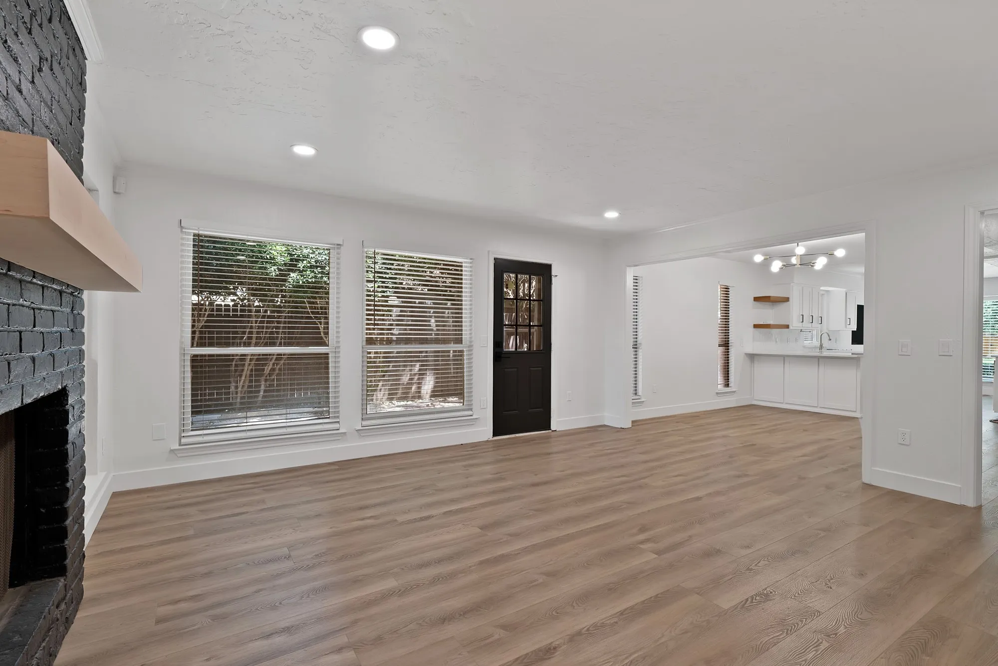 Spacious living area featuring light wood-finish flooring, a painted brick fireplace with a wood mantel, and recessed lighting