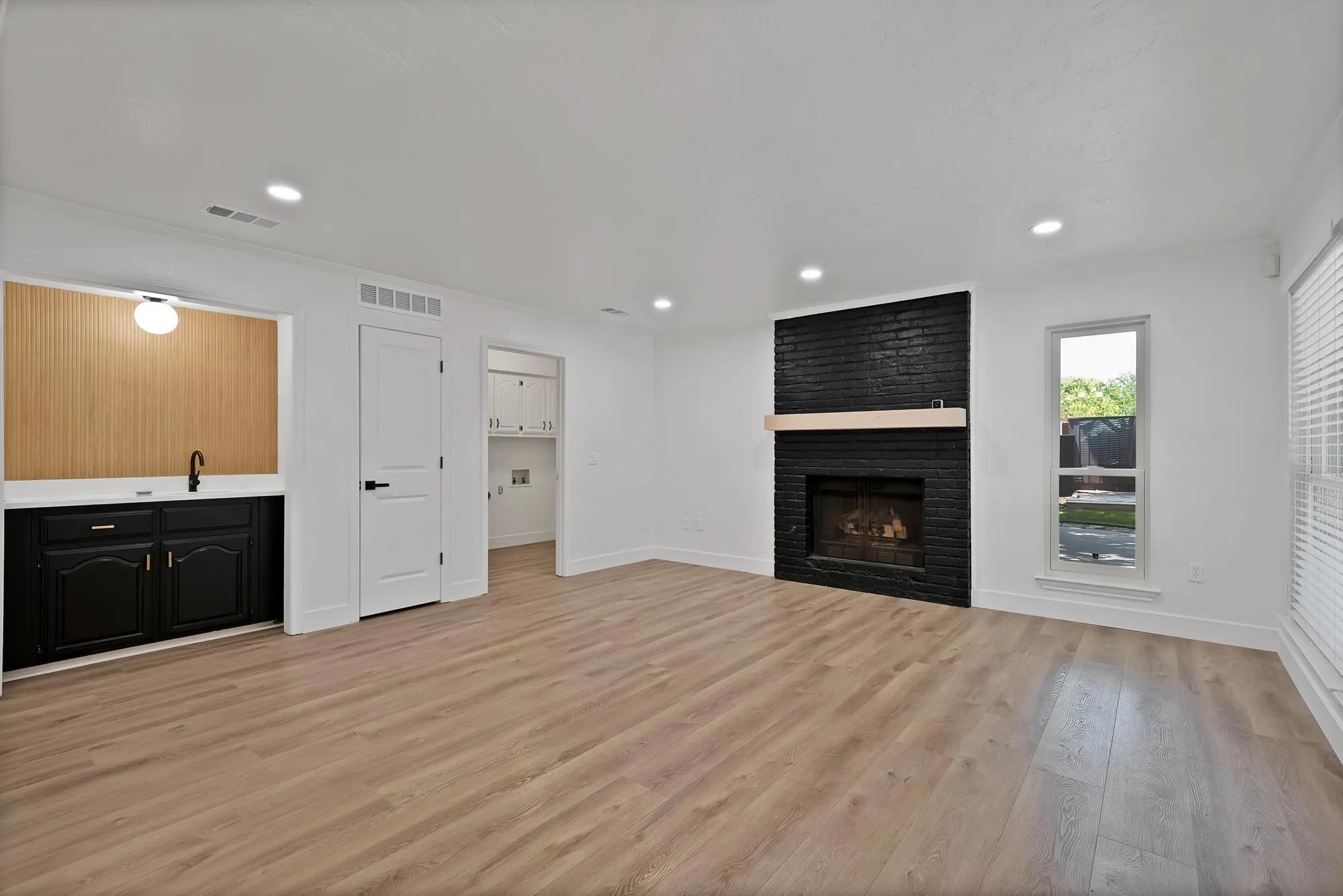 Living area featuring a black brick fireplace with a light wood mantel, wood-finish flooring, recessed lighting, and a built-in wet bar with black cabinetry and a light wood-paneled backsplash