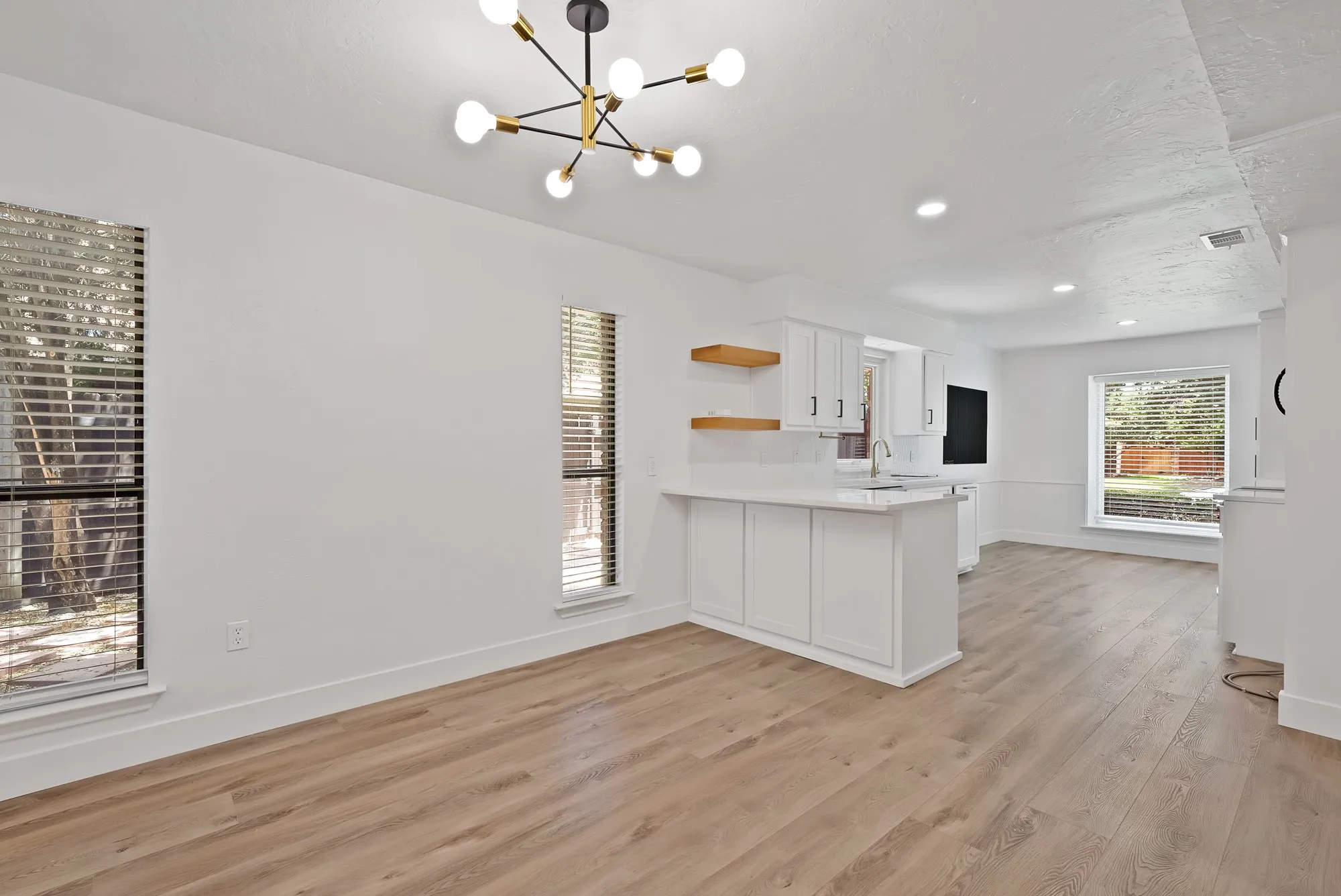 Open-concept living space featuring wood-finish flooring, a contemporary sputnik chandelier, multiple windows with horizontal blinds, and a built-in wet bar with white cabinetry and quartz countertop