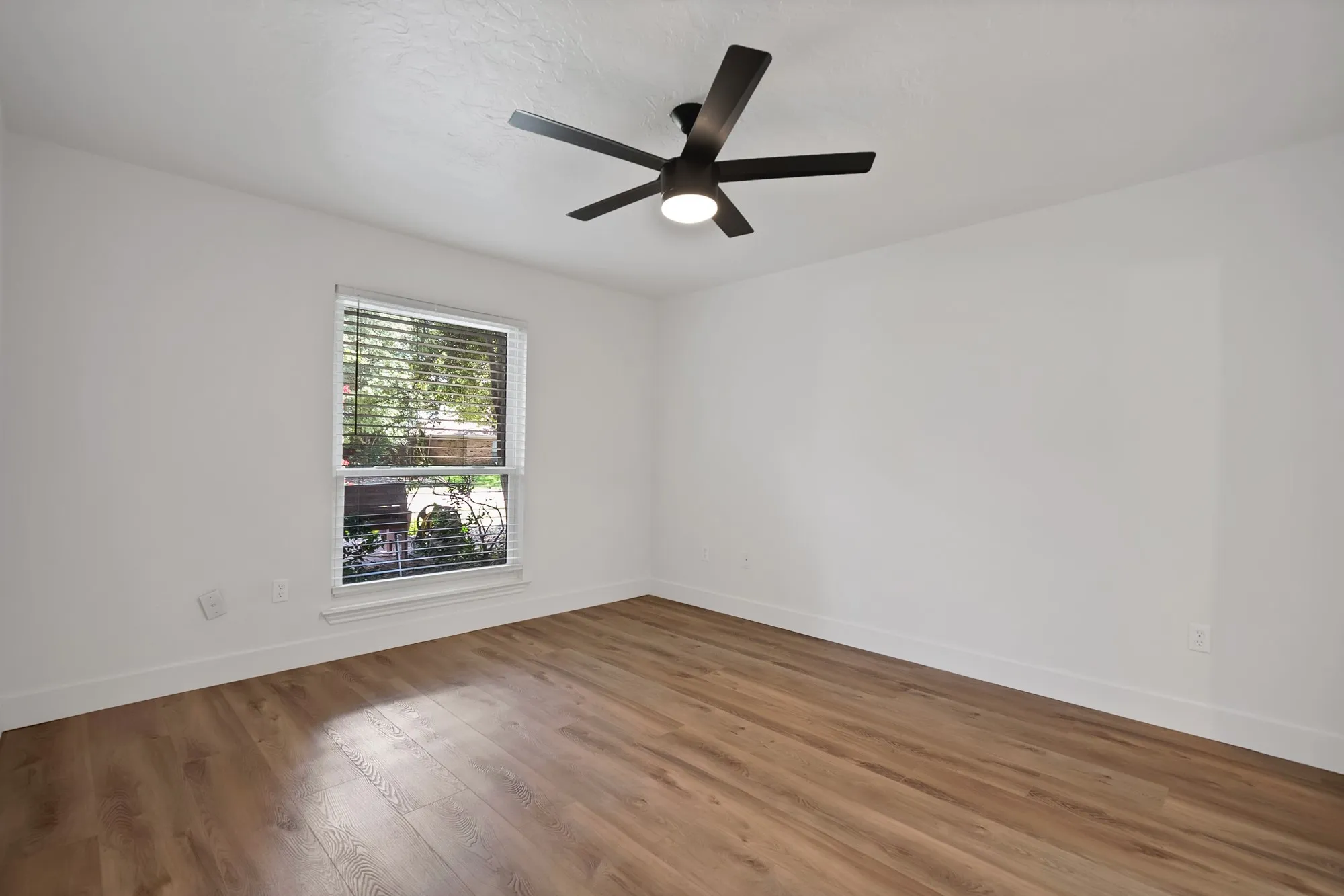 Spacious room featuring wood-finish flooring, a contemporary black ceiling fan with integrated lighting, and a window with horizontal blinds