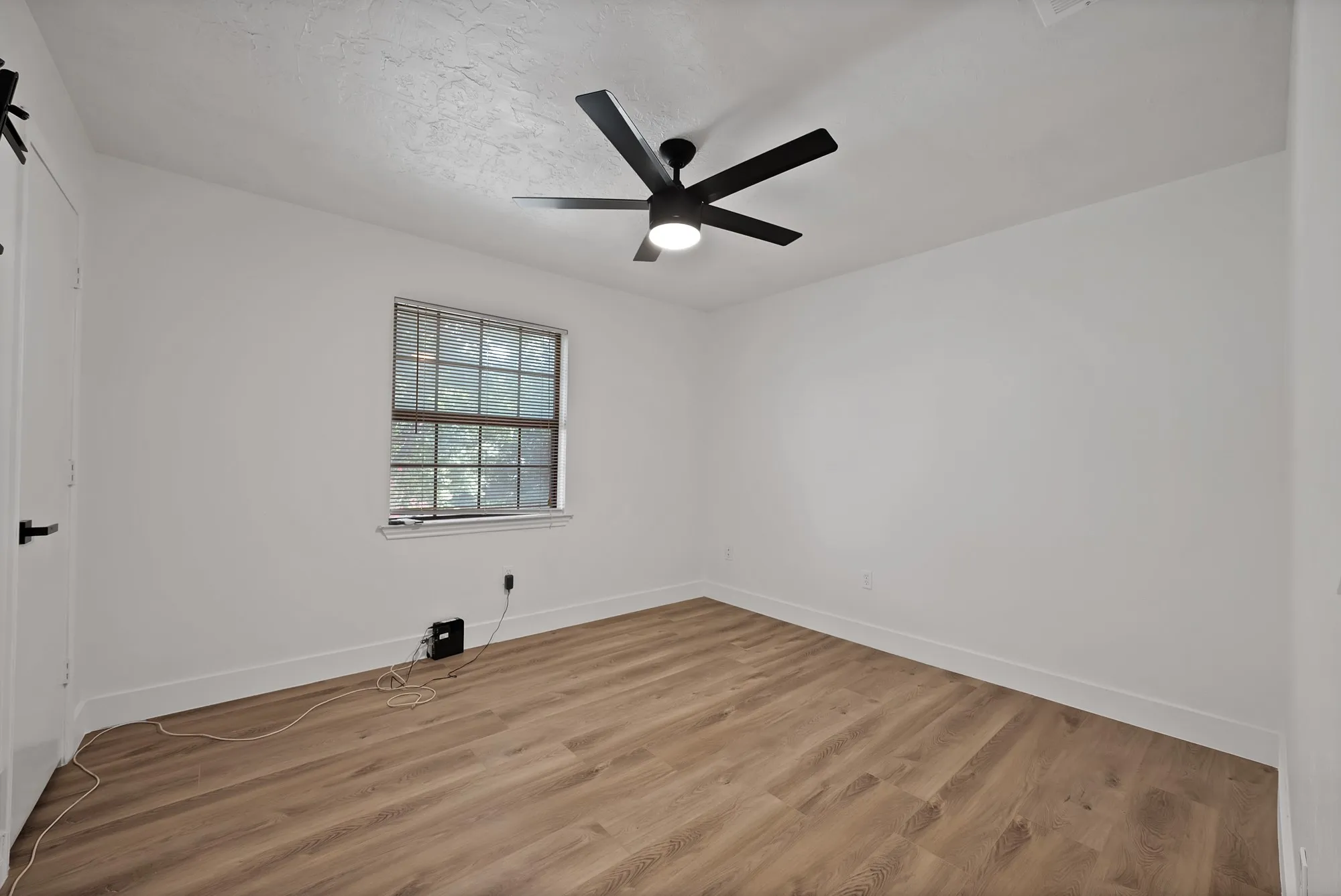 Bright room featuring wood-finish flooring, a contemporary ceiling fan with integrated lighting, a double-hung window, and a white interior door with matte black hardware