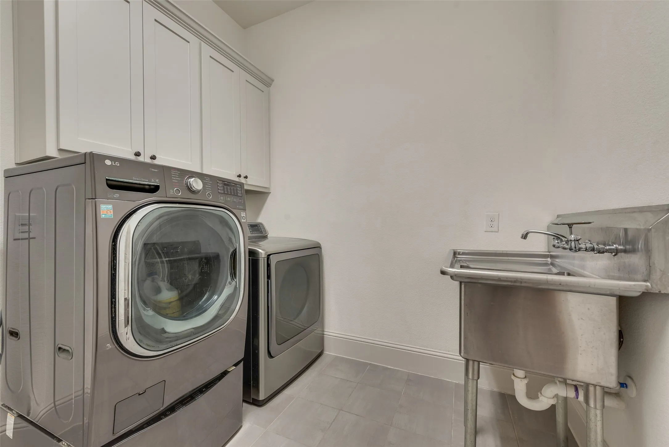 Utility room featuring upper cabinetry with dark hardware, a stainless steel utility sink with a wall-mounted faucet, and light-toned tile flooring