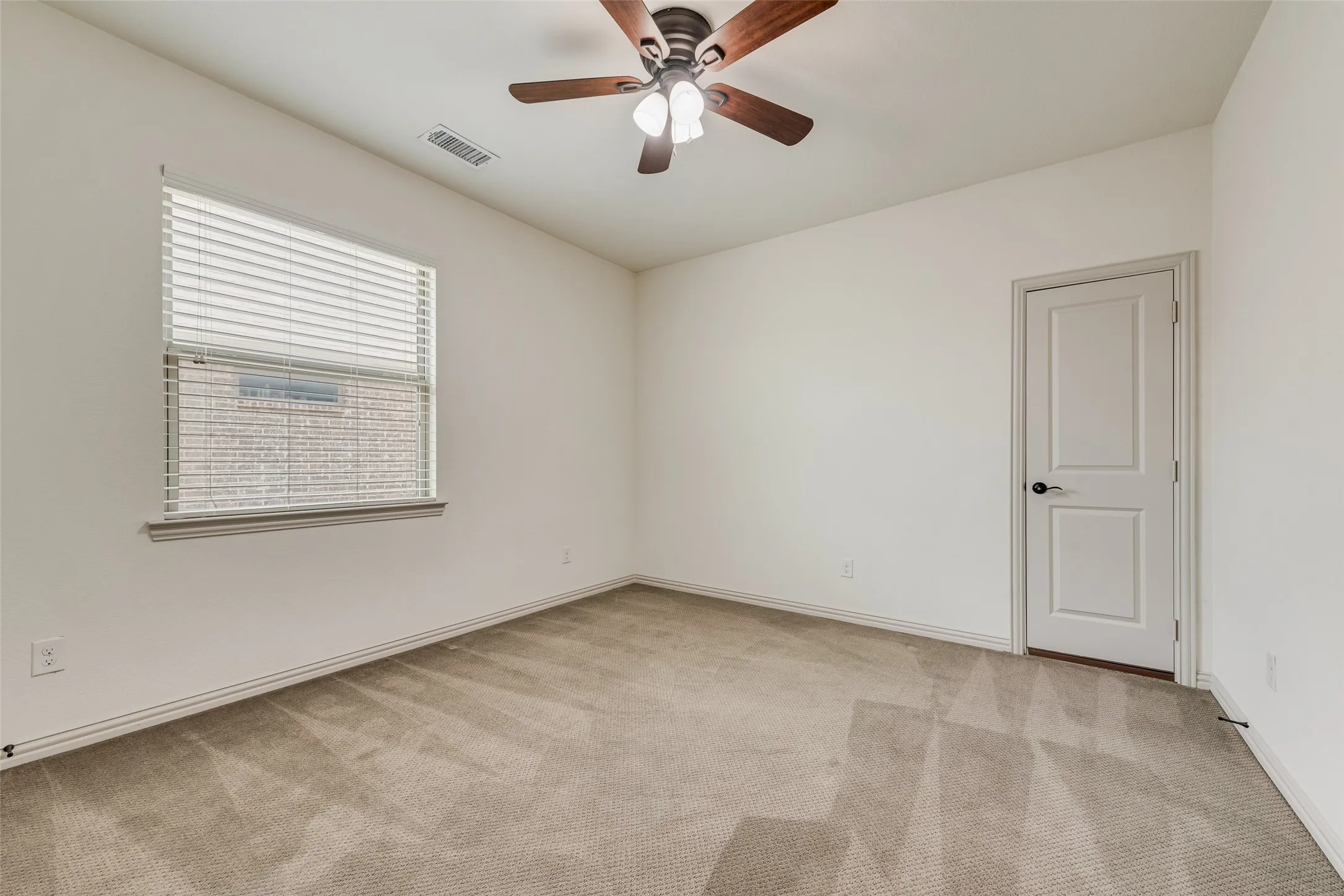 Spacious room featuring neutral carpet flooring, a ceiling fan with light fixture, a window with horizontal blinds, and a paneled door with dark hardware