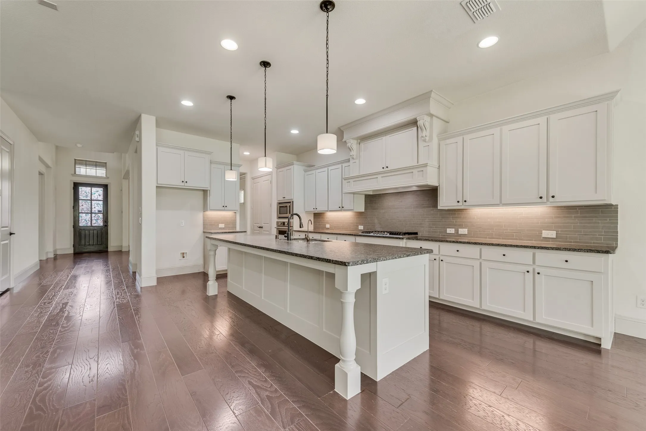 Open-concept kitchen featuring an oversized island with pendant lighting, white shaker cabinetry, a subway tile backsplash, and dark granite countertops