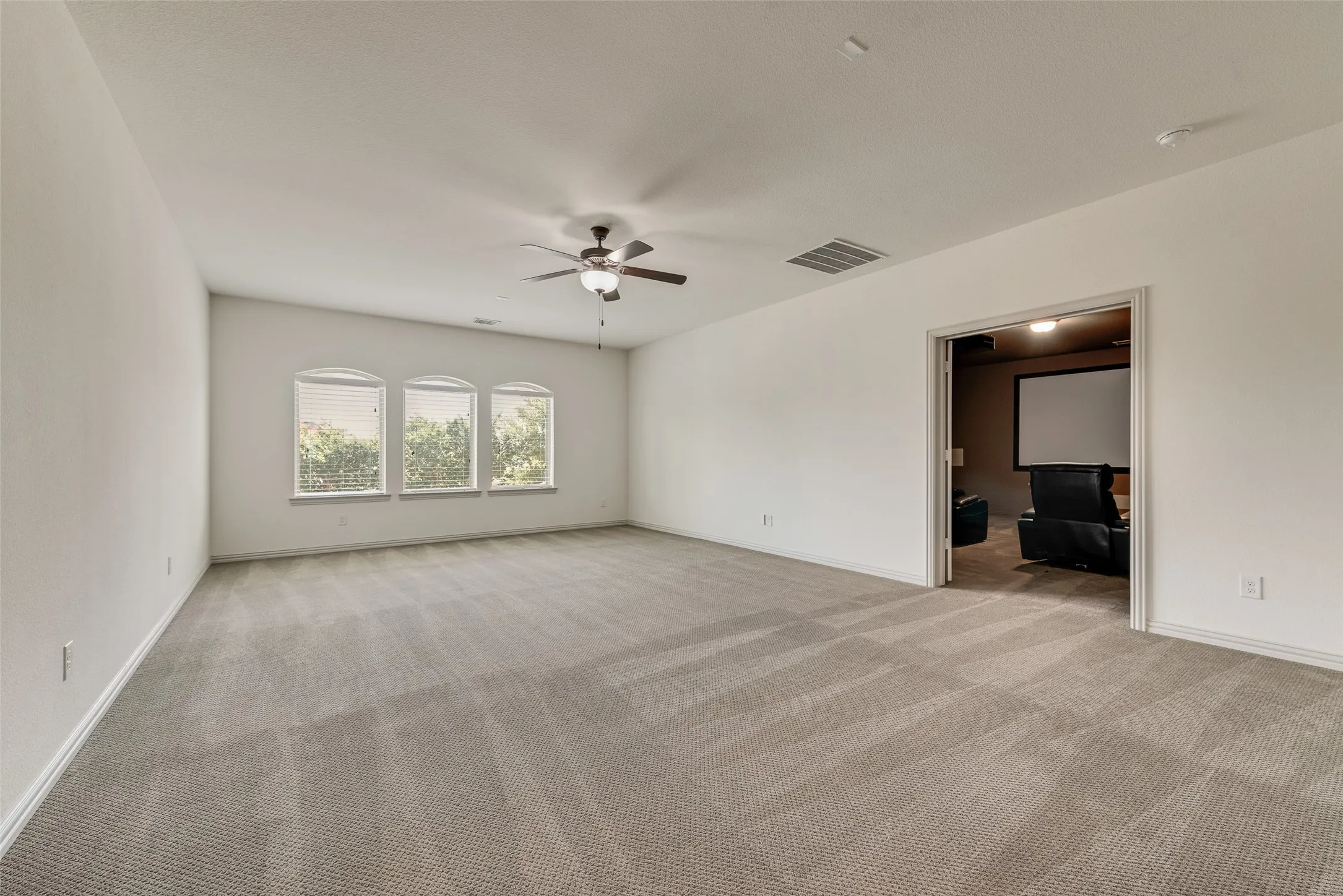 Spacious carpeted room featuring three arched windows with blinds, a ceiling fan with integrated lighting, and an open doorway leading to an additional room