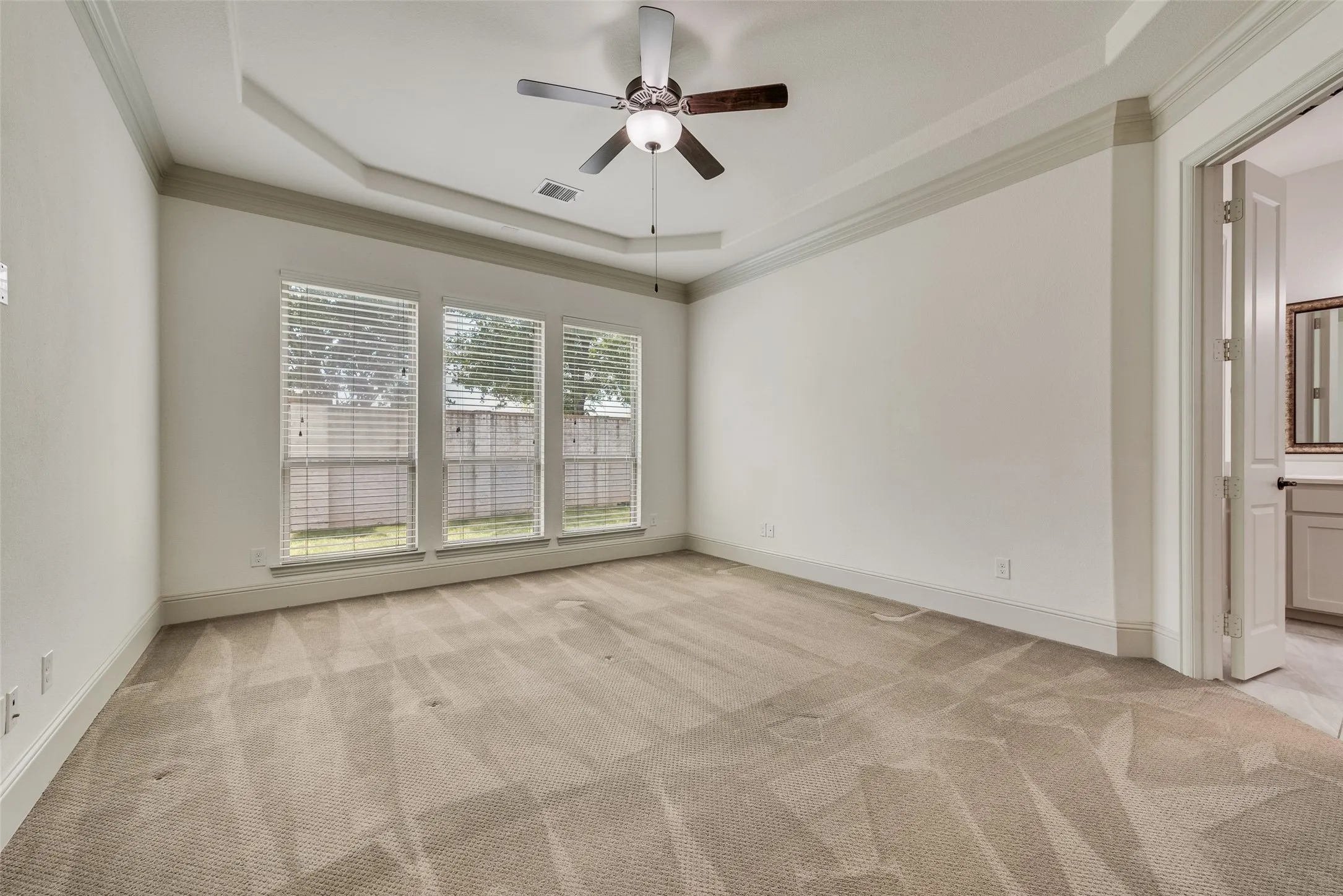 Spacious room featuring a tray ceiling with a ceiling fan, three large windows with blinds, plush carpeting, and crown molding