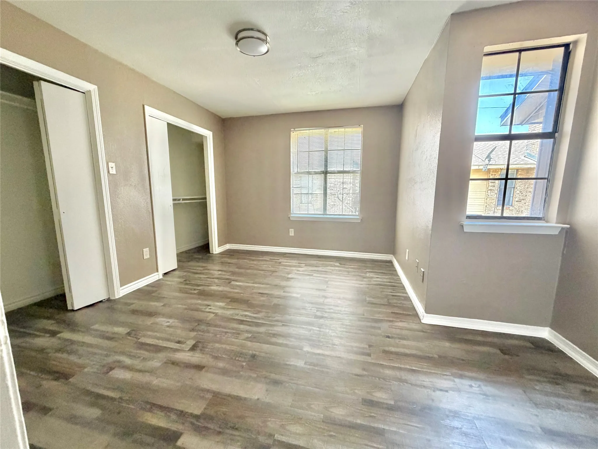 Spacious room featuring wood-finish flooring, two windows with white trim, a flush-mount ceiling light fixture, and two built-in closet spaces with bi-fold doors