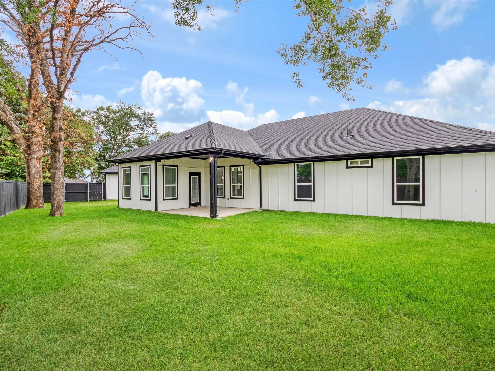 Expansive rear yard featuring a covered patio with concrete flooring, vertical white siding, black-framed windows, and a black shingle roof