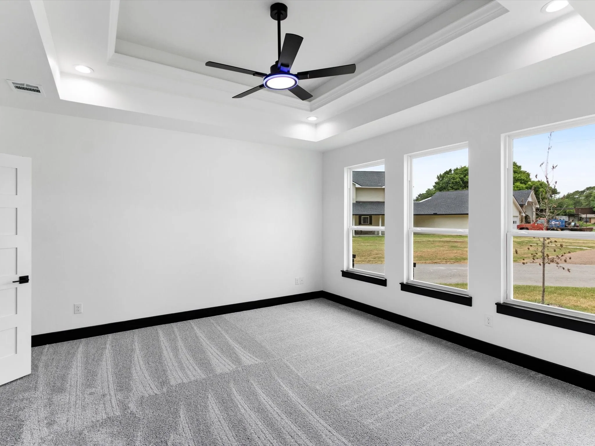 Spacious room featuring a tray ceiling with recessed lighting, black modern ceiling fan, three pane windows, white walls, and gray patterned carpeting