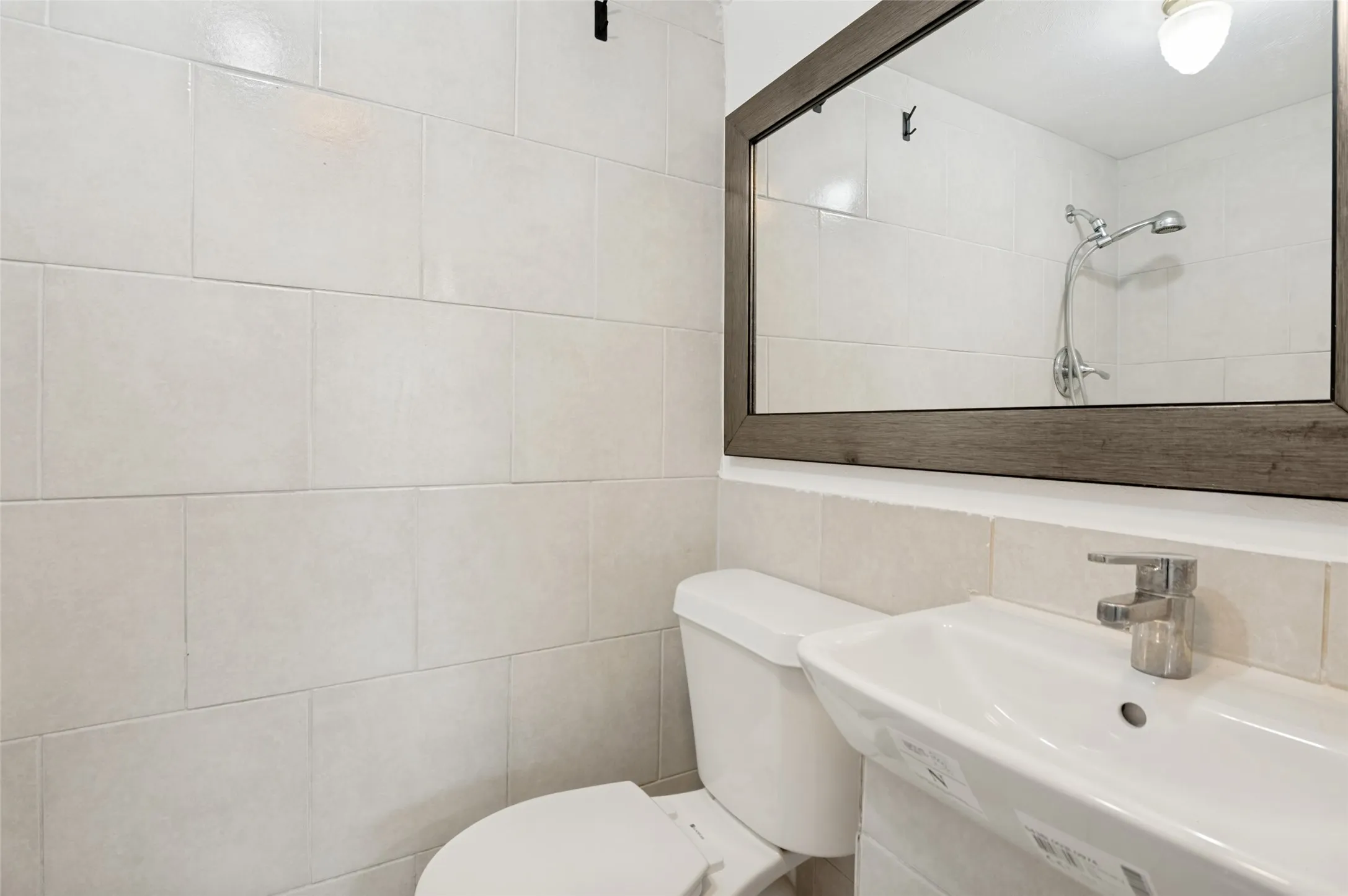 Bathroom featuring light-toned large format wall tiling, a rectangular vessel sink with a chrome faucet, a white toilet, and a framed mirror