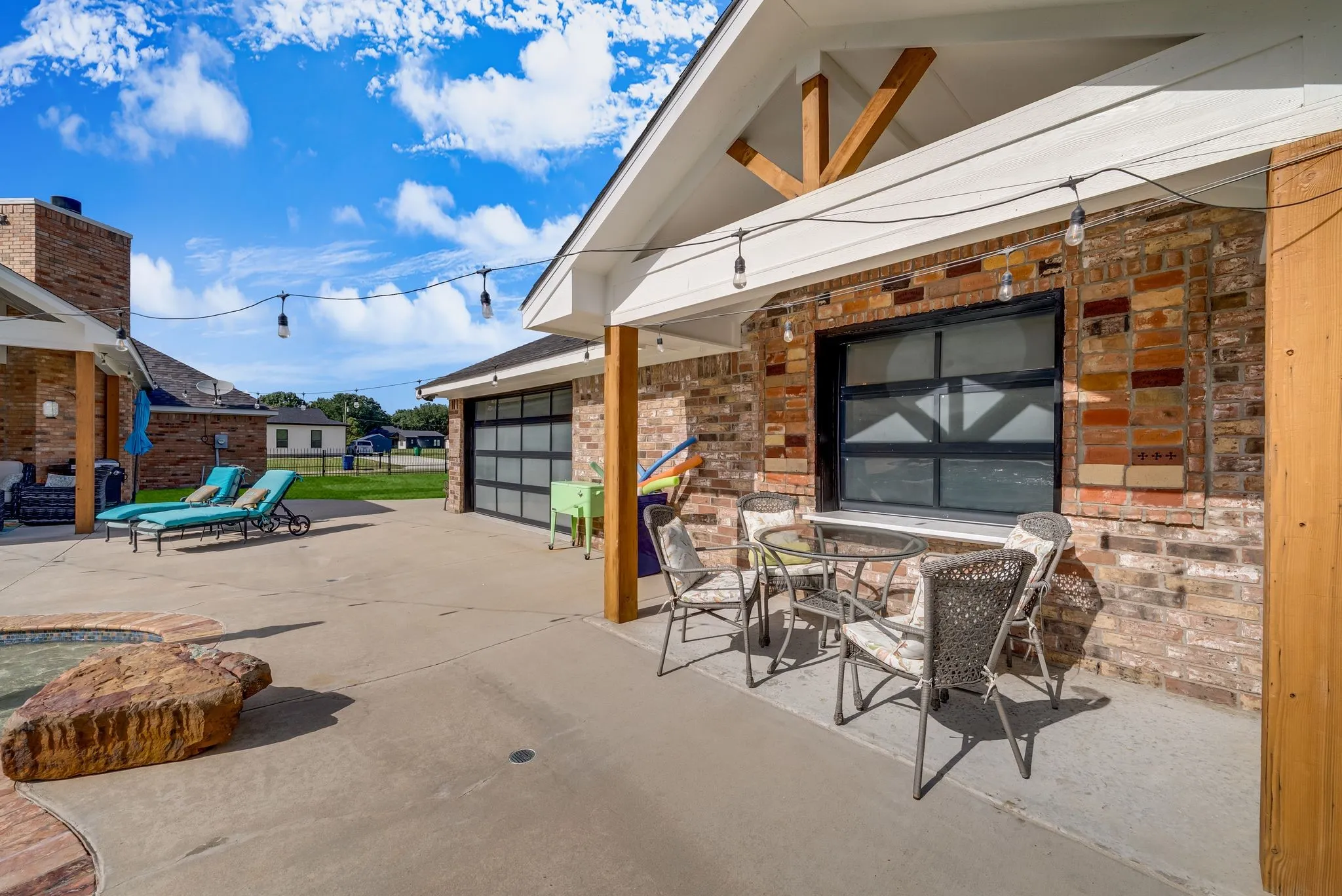 Expansive concrete patio area featuring string lights, a brick-clad exterior, a modern black garage door, and exposed timber ceiling beams