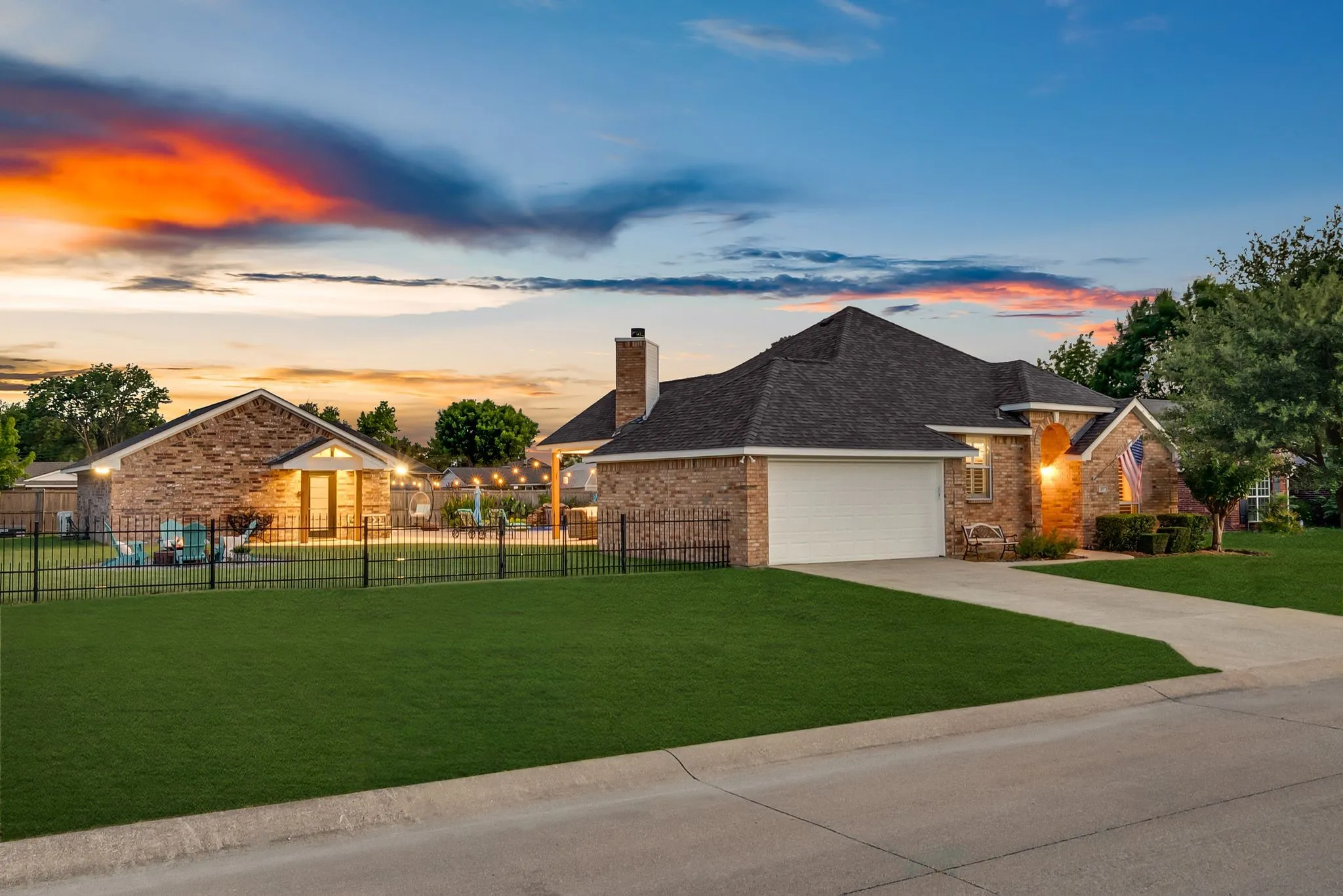 Brick exterior featuring an attached two-car garage, a separate detached brick structure, a black wrought iron fence, a concrete driveway, and mature landscaping