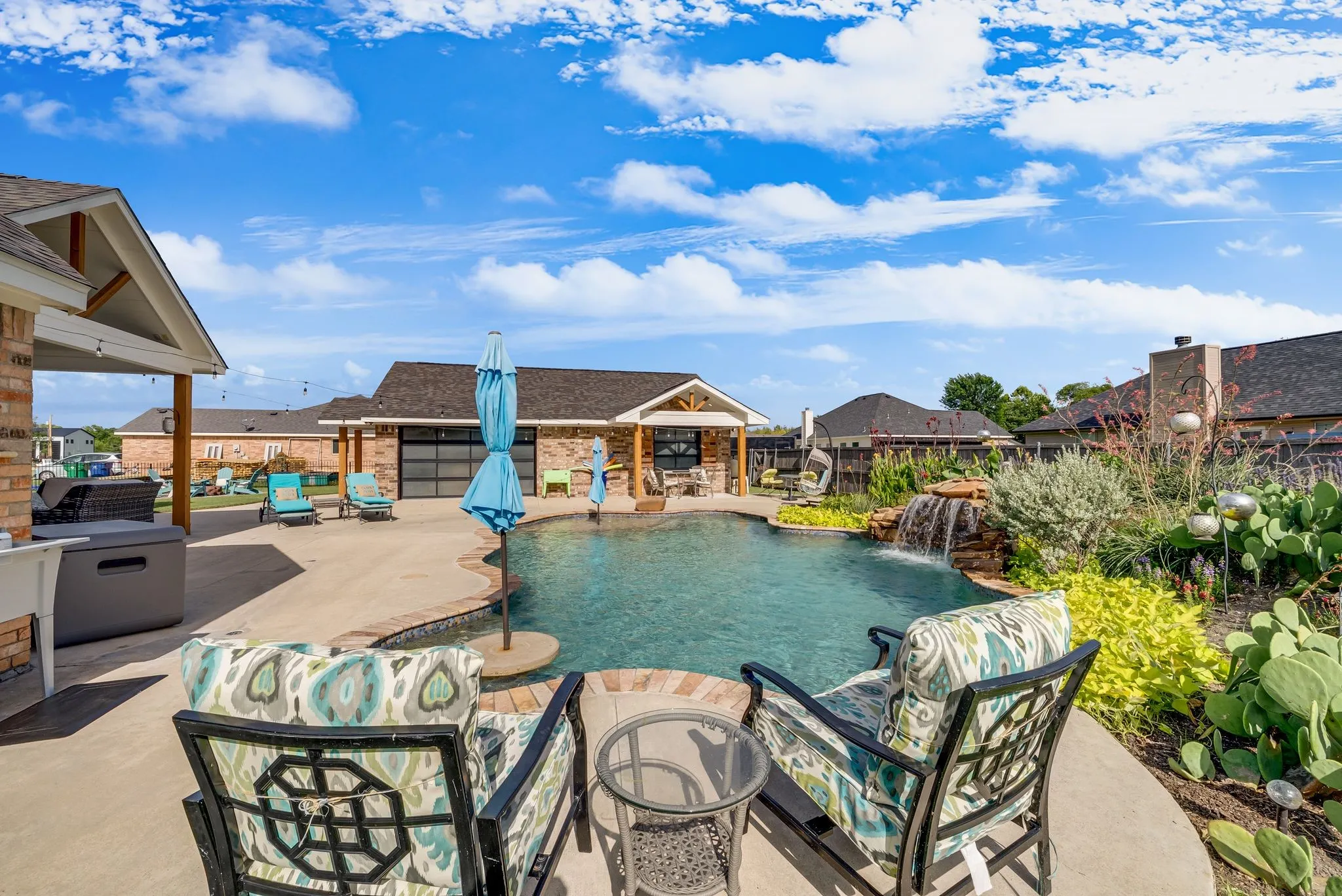Outdoor concrete patio featuring a built-in swimming pool with a stone waterfall, a covered patio area with exposed beams, and mature landscaping with various plants
