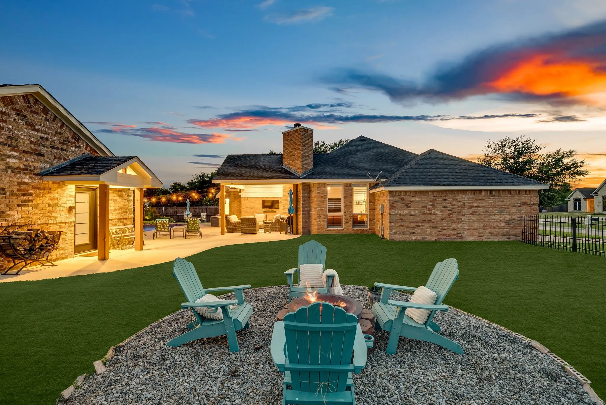 Expansive backyard featuring a central gravel fire pit area with Adirondack chairs, a manicured lawn, and a brick-built home with a covered​​‌​​​​‌​​‌‌​‌‌​​​‌‌​‌​‌​‌​​​‌​​ patio