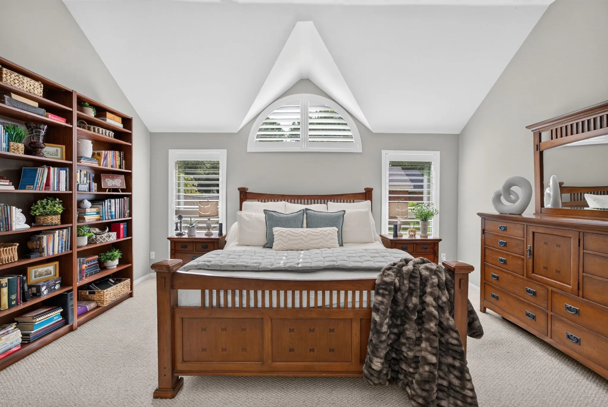 Vaulted ceiling bedroom with light gray walls, plush carpet, two full-height built-in bookcases, two rectangular windows with plantation shutters, and an arched transom window with plantation shutters