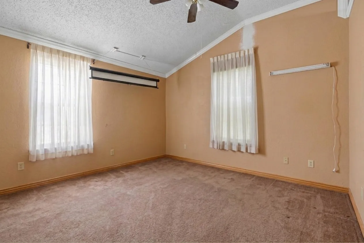 Carpeted interior space featuring a vaulted ceiling with a ceiling fan, crown molding, and two windows with sheer curtains