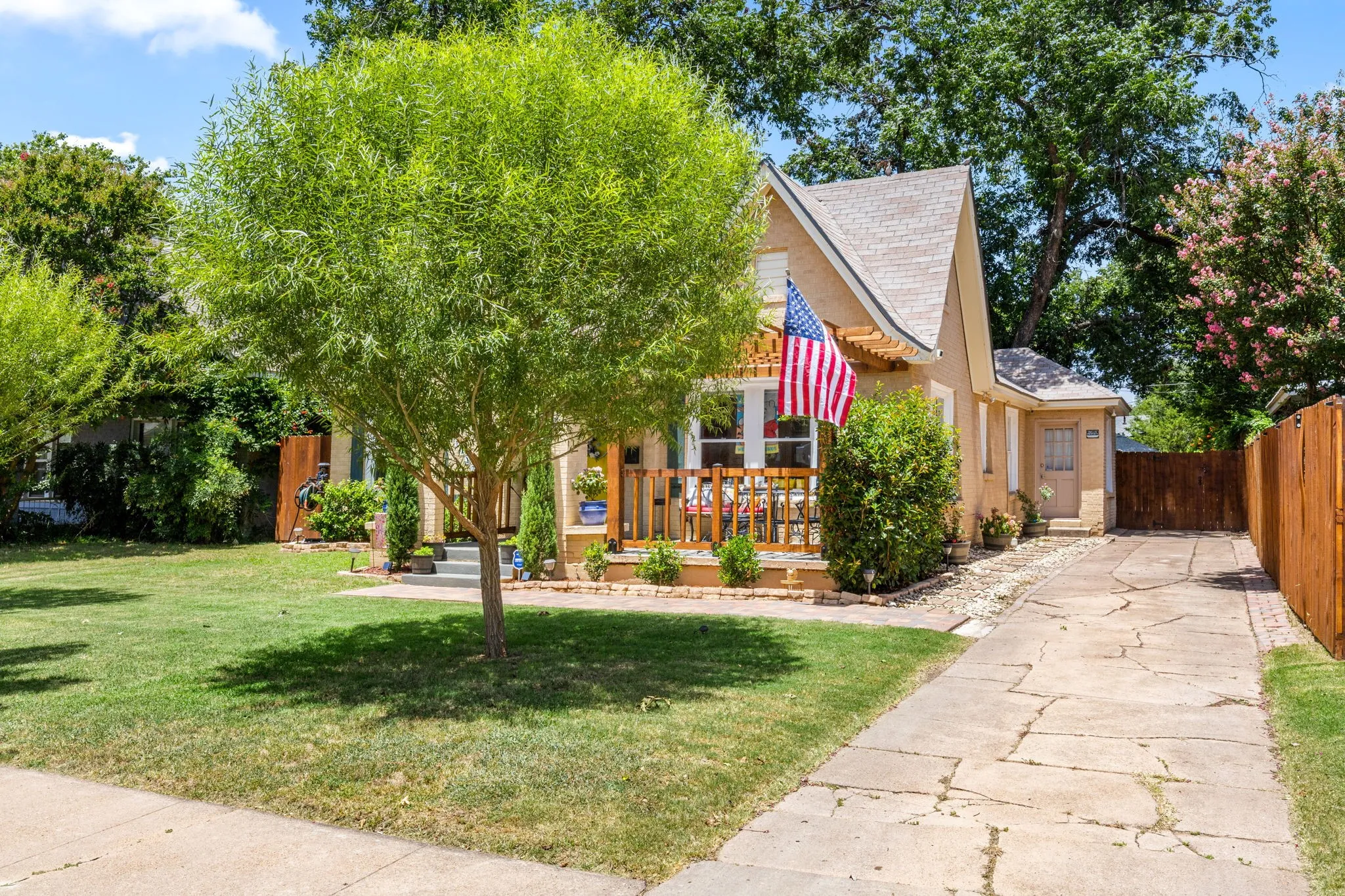 Front exterior featuring a covered porch, gabled roofline, mature trees, concrete driveway and wooden privacy fence