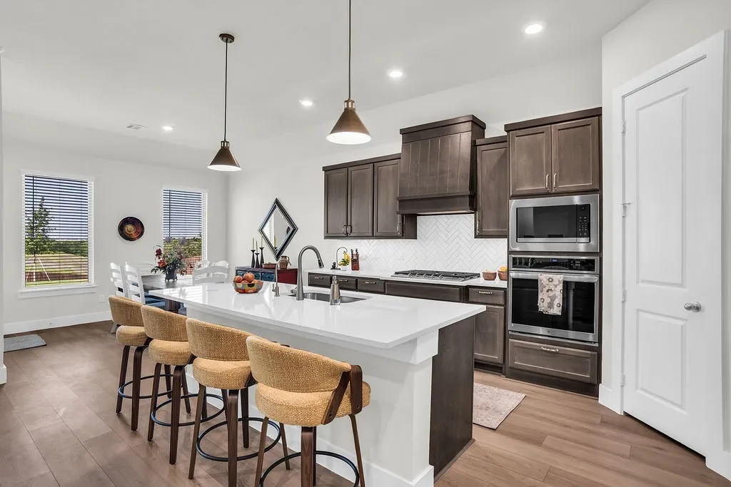 Kitchen featuring a substantial island with breakfast bar, undermount sink with gooseneck brushed nickel faucet, wood-finish flooring, dark wood cabinetry, a white herringbone tile backsplash, walk-in pantry and stainless steel appliances including a propane gas cooktop, electric built-in oven and microwave open to living and breakfast rooms