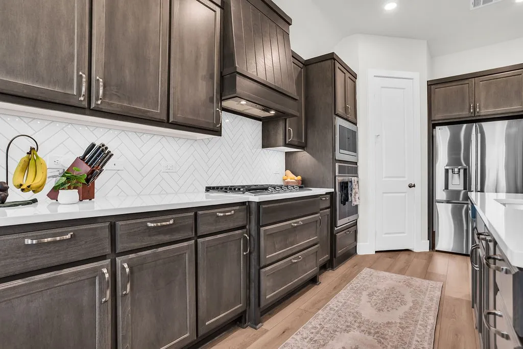 Kitchen featuring a substantial island with breakfast bar, undermount sink with gooseneck brushed nickel faucet, wood-finish flooring, dark wood cabinetry, a white herringbone tile backsplash, walk-in pantry and stainless steel appliances including a propane gas cooktop, electric built-in oven and microwave open to living and breakfast rooms