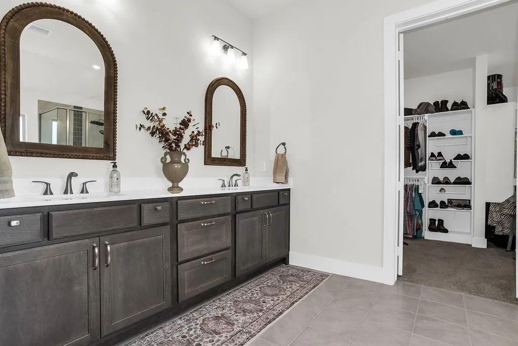 Main ensuite bath features dual vanity with wood-finish cabinetry, brush bronze faucets,  glass-enclosed steam shower with a tiled surround and vertical accent, a freestanding soaking tub ceramic tile floors and spacious walk-in closet with built-in shelving.