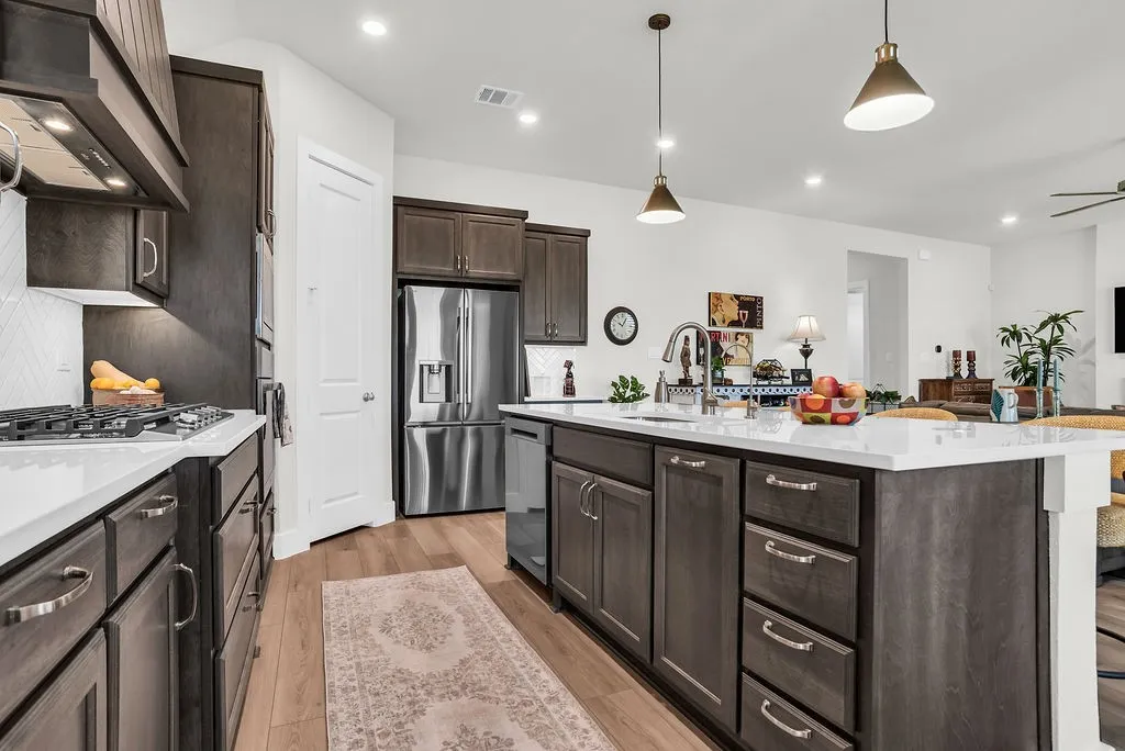 Kitchen featuring a substantial island with breakfast bar, undermount sink with gooseneck brushed nickel faucet, wood-finish flooring, dark wood cabinetry, a white herringbone tile backsplash, walk-in pantry and stainless steel appliances including a propane gas cooktop, electric built-in oven and microwave open to living and breakfast rooms