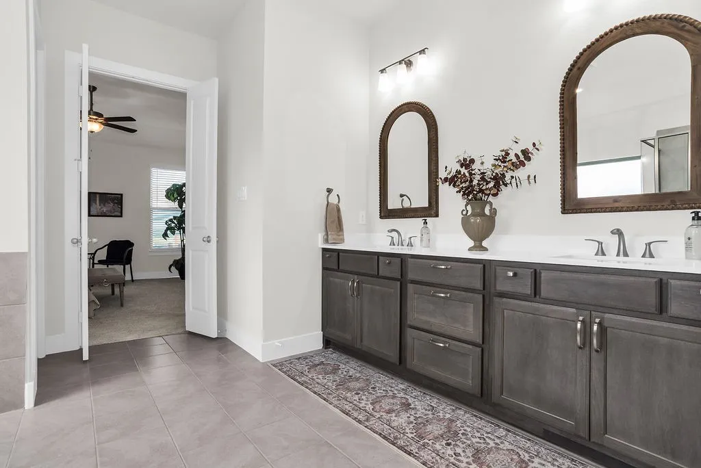 Main ensuite bath features dual vanity with wood-finish cabinetry, brush bronze faucets,  glass-enclosed steam shower with a tiled surround and vertical accent, a freestanding soaking tub ceramic tile floors and spacious walk-in closet with built-in shelving.