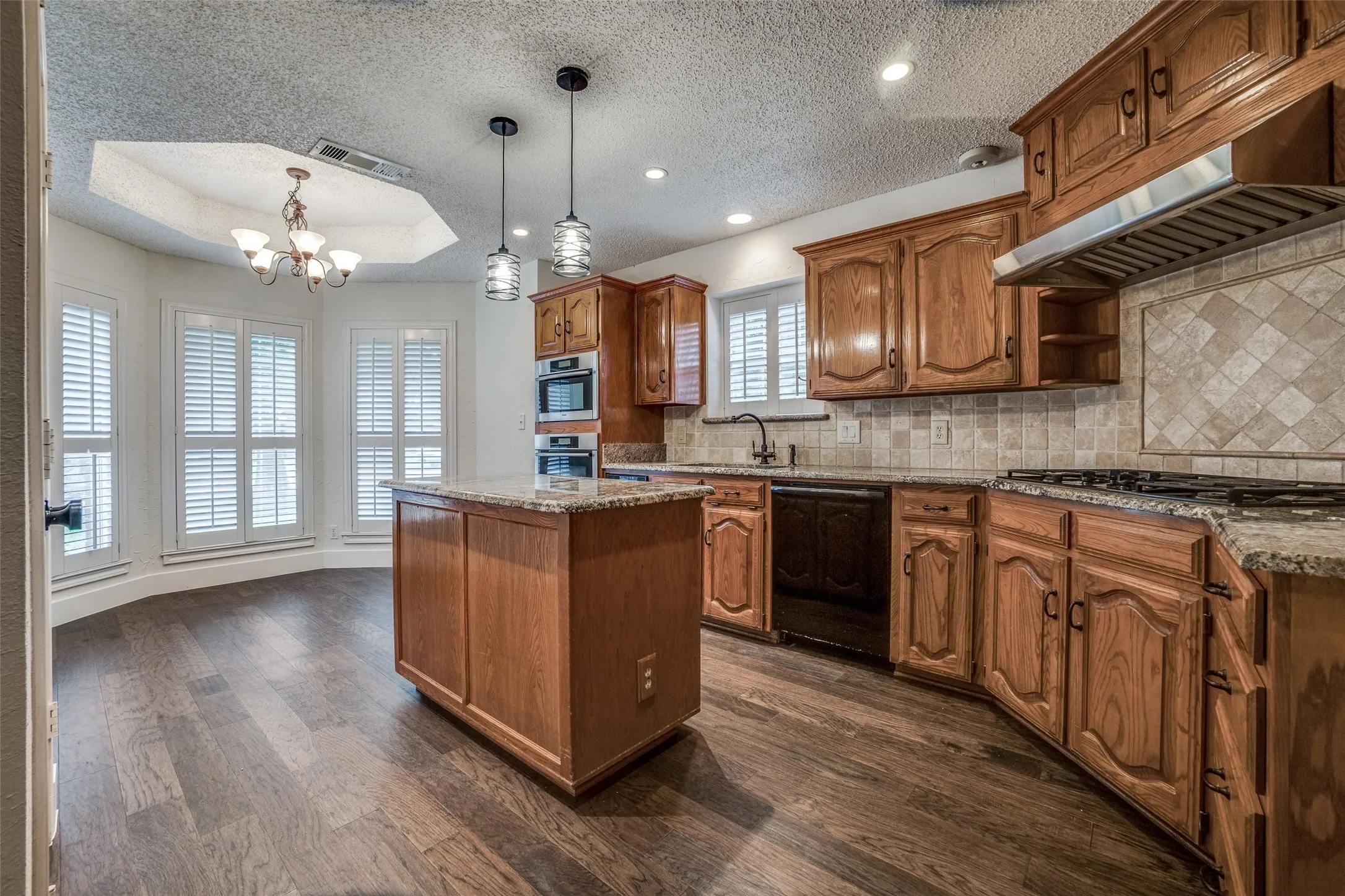 Kitchen featuring wood-finish flooring, a central island with a stone countertop, wood cabinetry, built-in double ovens, and a gas range with a stainless steel vent hood