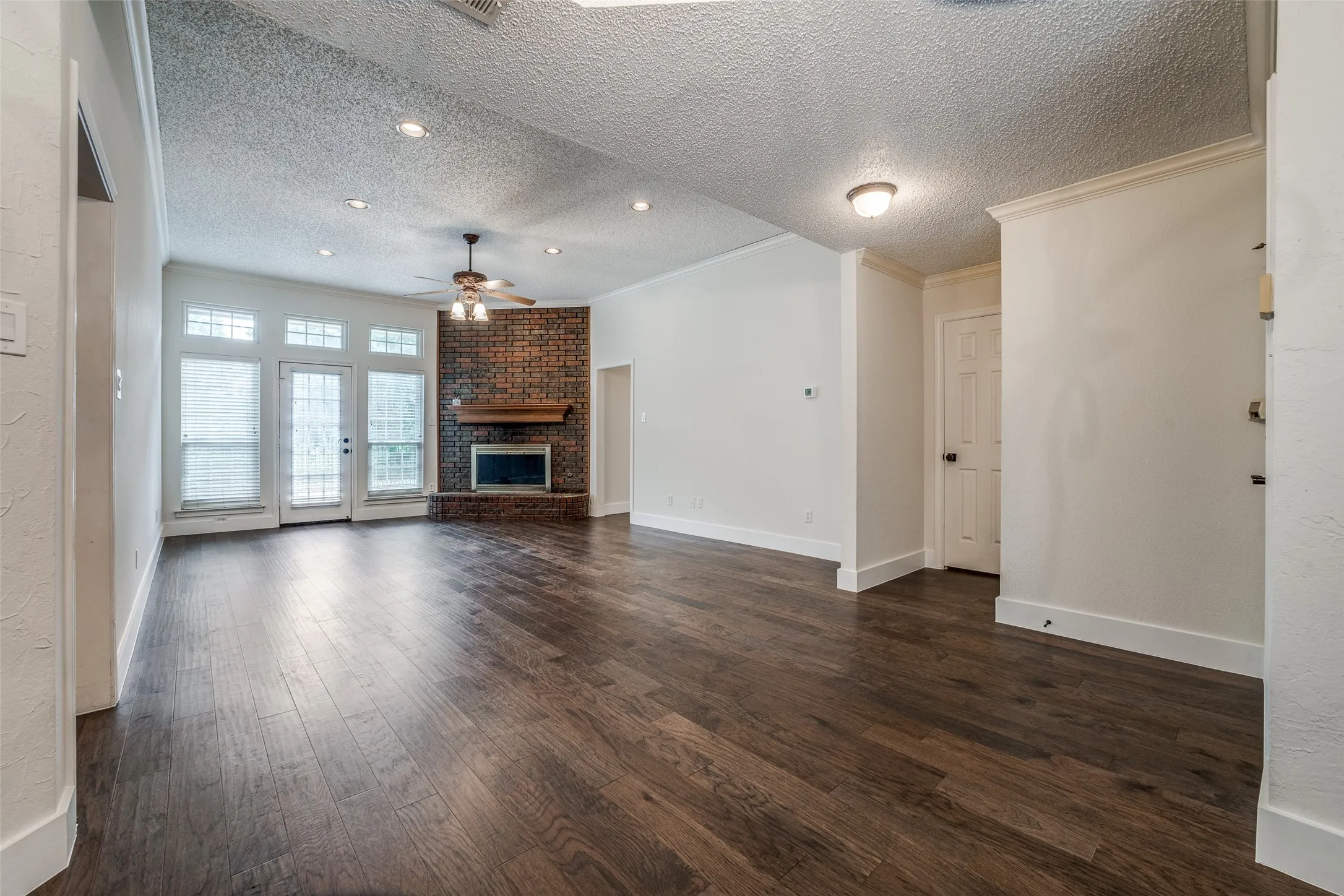 Spacious living area featuring wood-finish flooring, a brick fireplace with a wood mantle, and a vaulted ceiling with recessed lighting