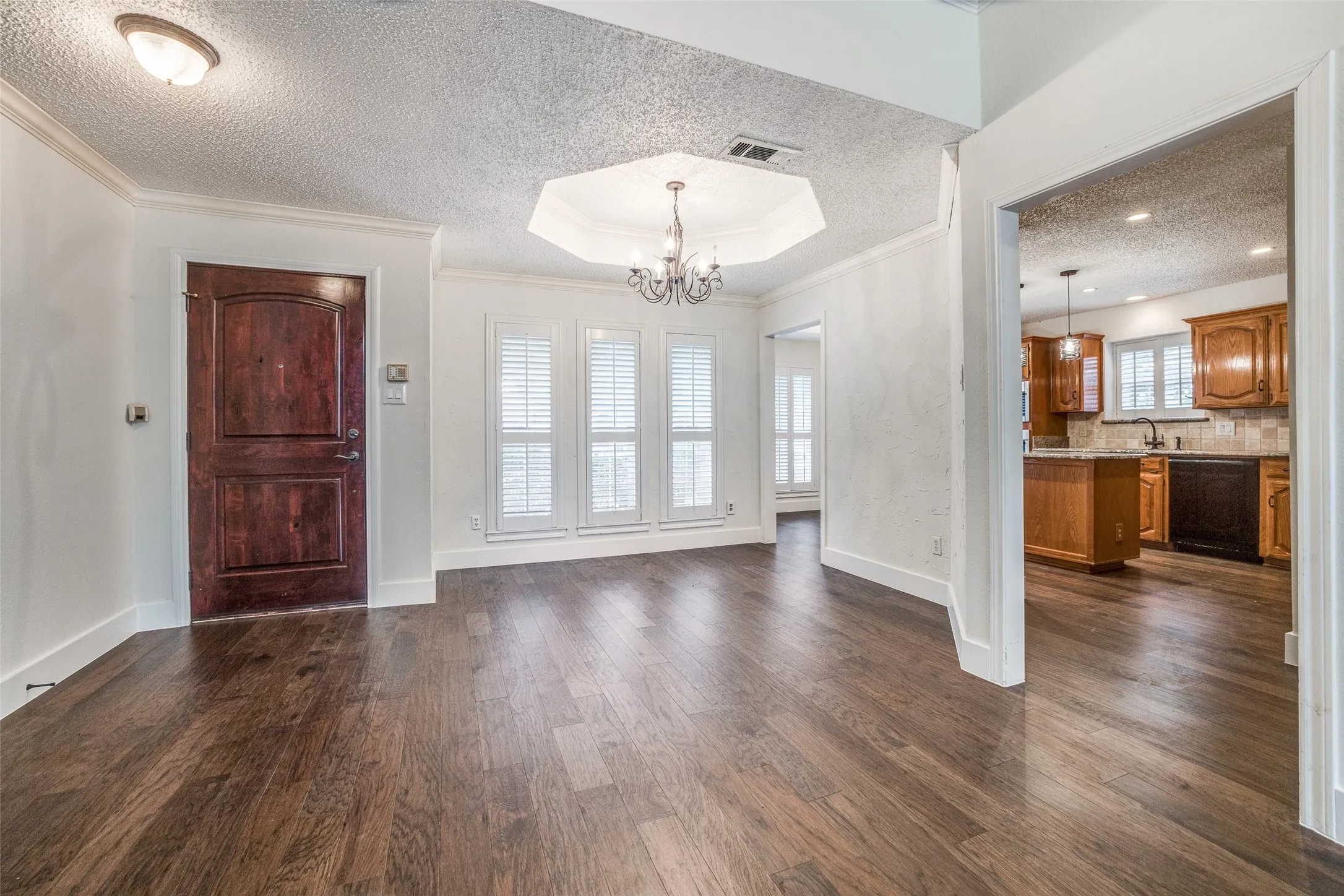 Warm wood-finish flooring throughout, featuring a rich dark wood entry door, multiple windows with plantation shutters, an octagonal tray ceiling with a chandelier, and crown molding