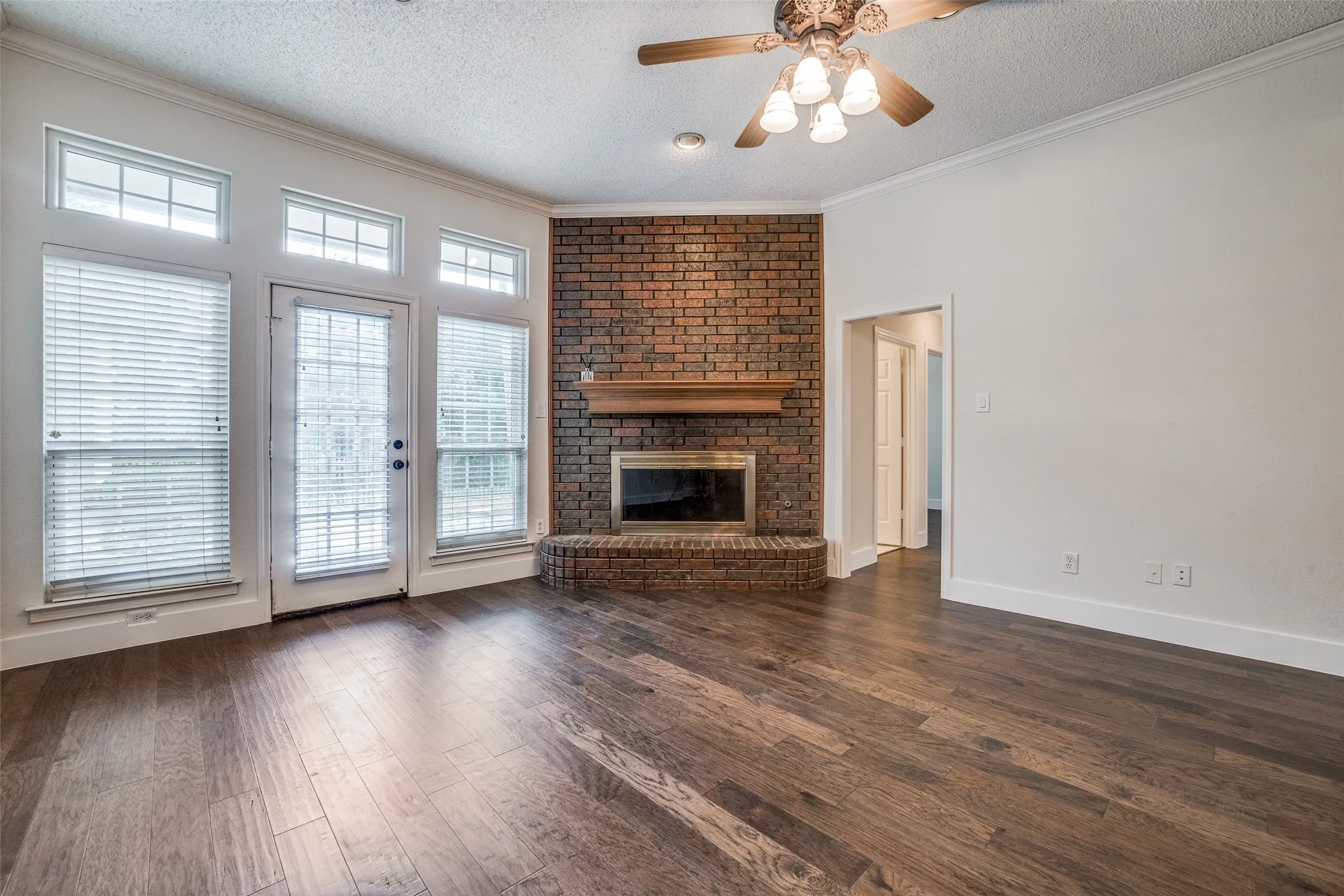 Spacious living area featuring wood-finish flooring, a brick fireplace with a wood mantle, and a glass-paneled door flanked by two large windows
