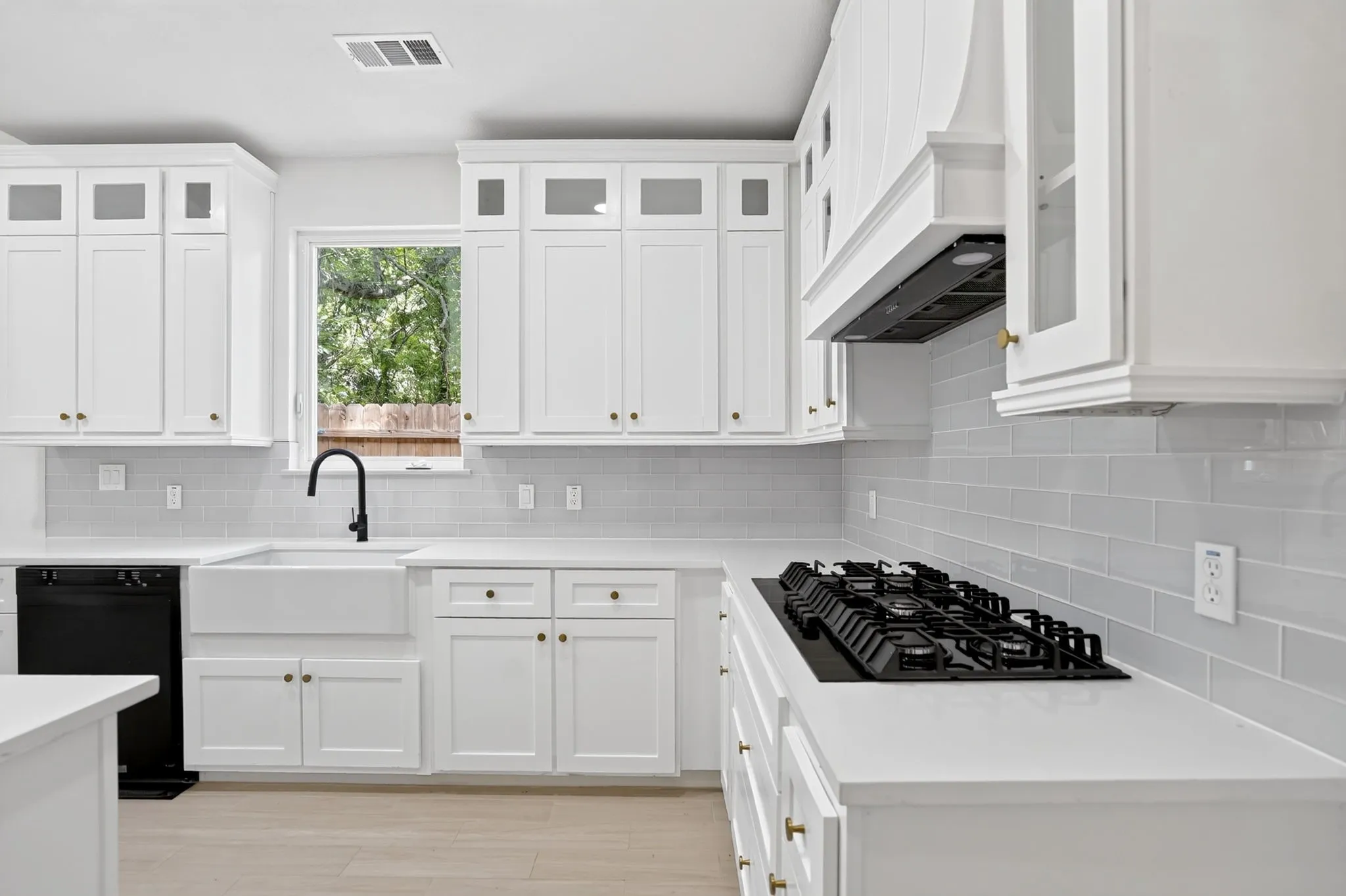 White shaker cabinetry with brass hardware, a white apron-front sink with matte black faucet, light grey subway tile backsplash, white countertops, and a built-in gas cooktop with matching range hood