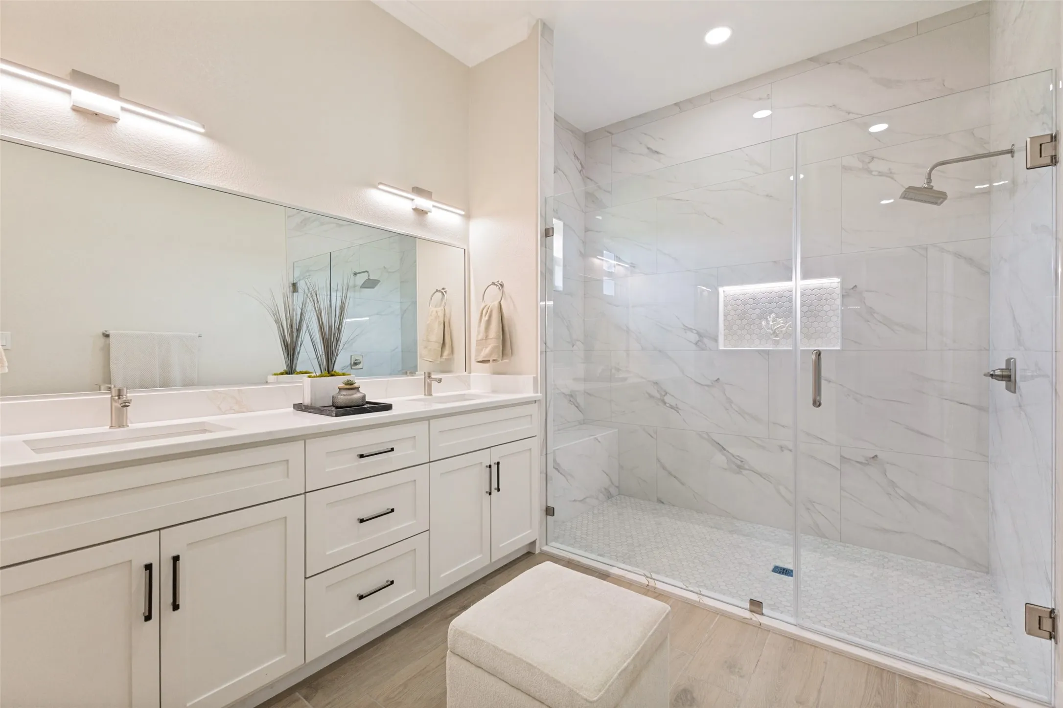 Spacious primary bathroom featuring a dual vanity with white cabinetry and quartz countertops, an expansive mirror, and a glass-enclosed shower with large format marble-look tile walls and a hexagonal tile floor