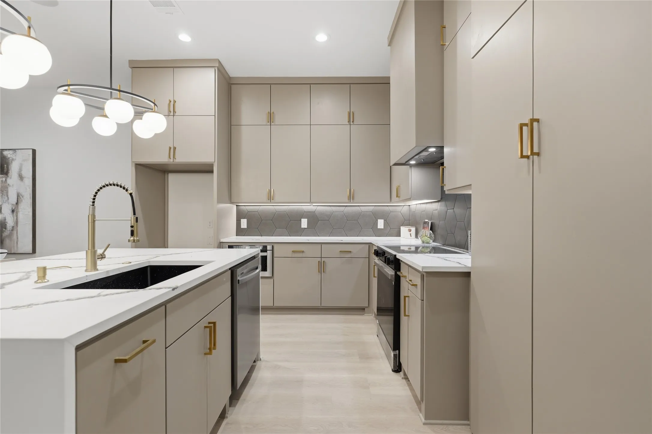 Open-concept kitchen featuring light-toned cabinetry with gold-finish hardware, a large island with a black sink and gold-finish faucet, white stone countertops, and a gray hexagonal tile backsplash