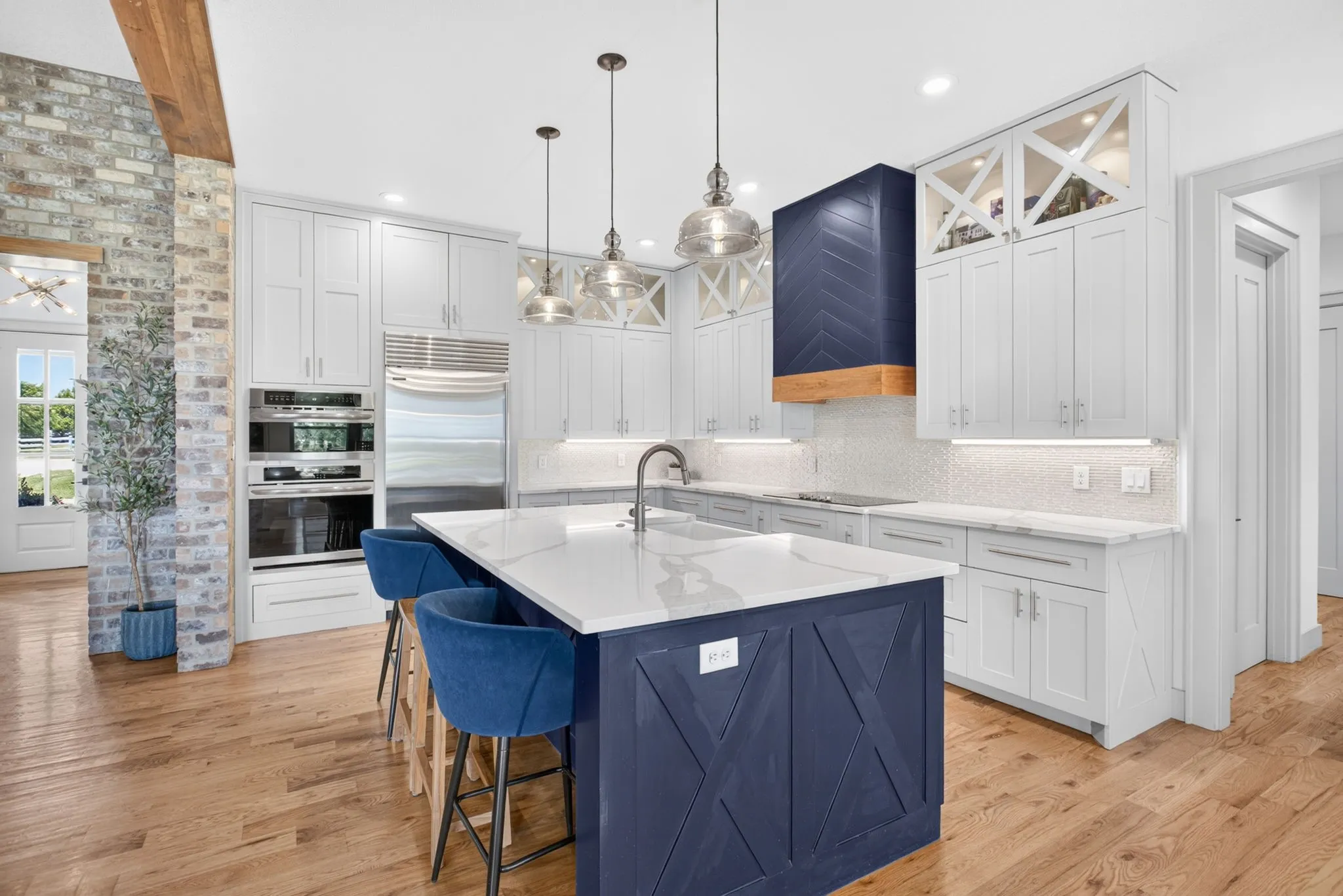 Gourmet kitchen featuring a large central island with a quartz countertop and navy blue base cabinetry, light wood-finish flooring, white perimeter cabinetry, stainless steel built-in appliances, and a unique range hood with a chevron pattern