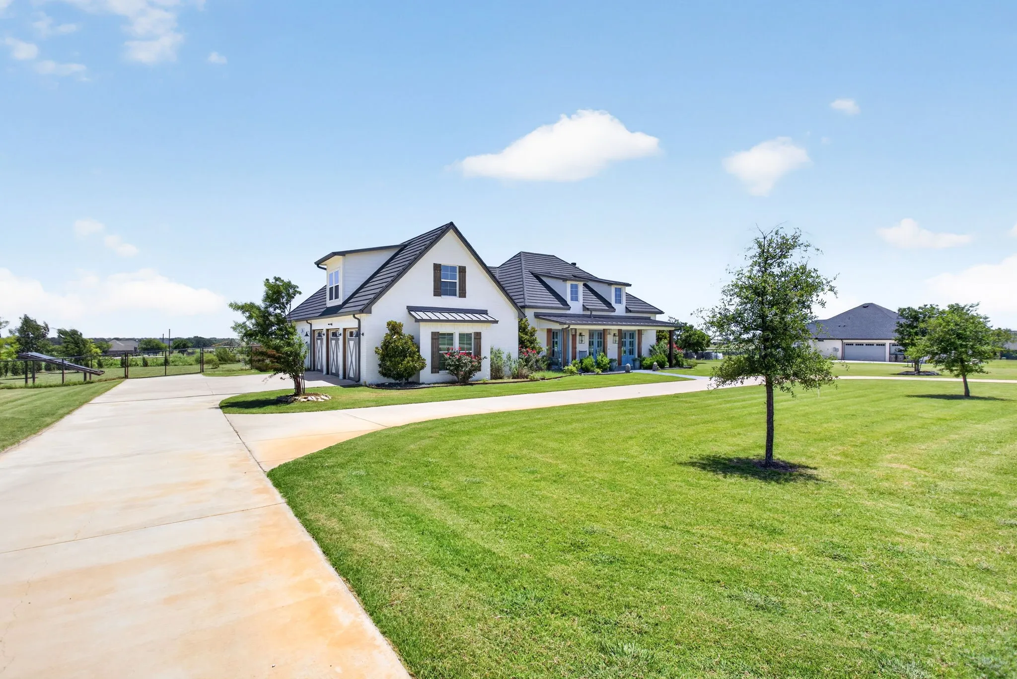 White exterior with dark trim and shutters, featuring a prominent front porch with blue double doors, a large green lawn, an expansive concrete driveway, and a three-car garage