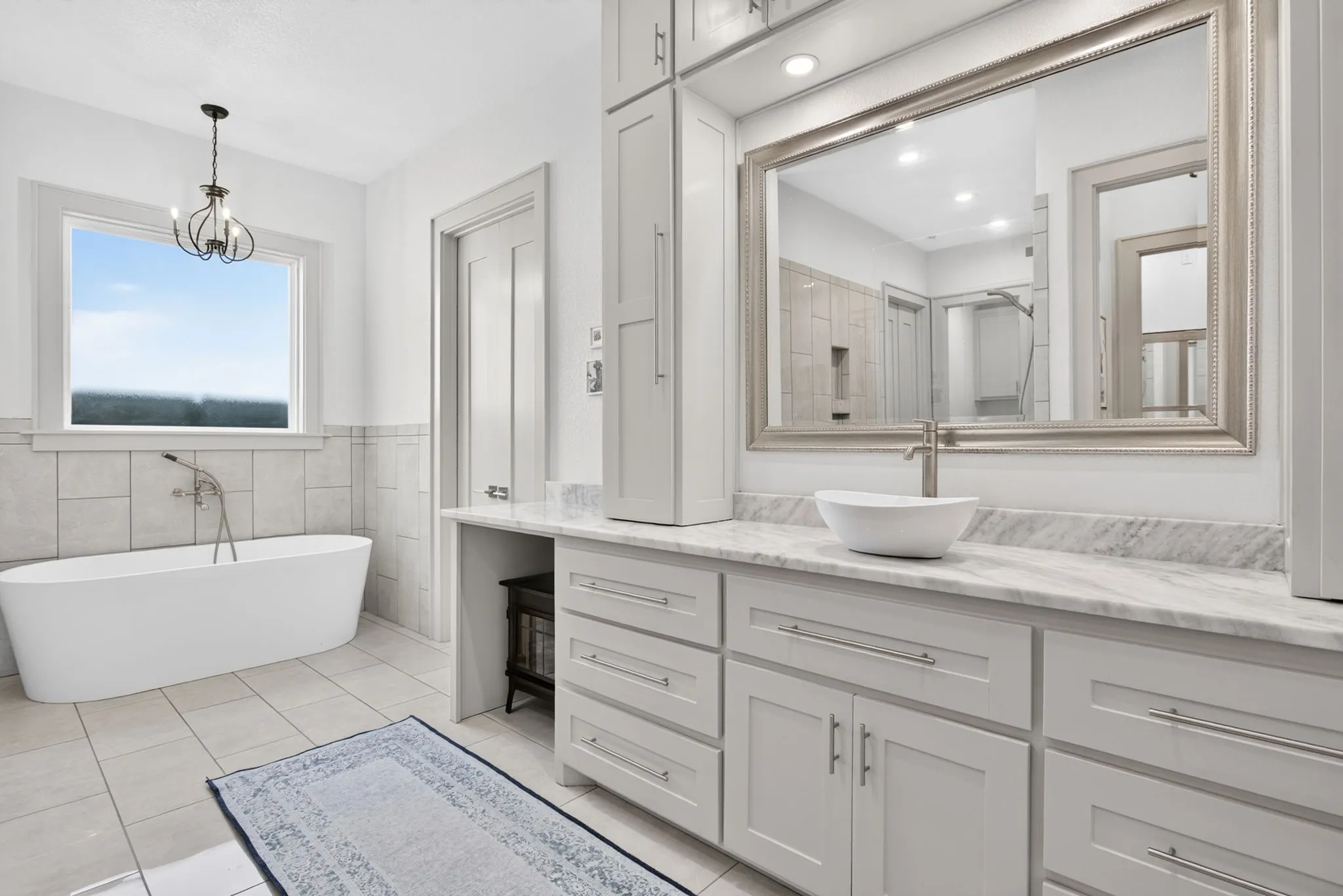 Bathroom featuring a freestanding soaking tub with floor-mounted tub filler, light-toned tile wainscoting, and a large vanity with a vessel sink, stone countertop, and extensive cabinetry