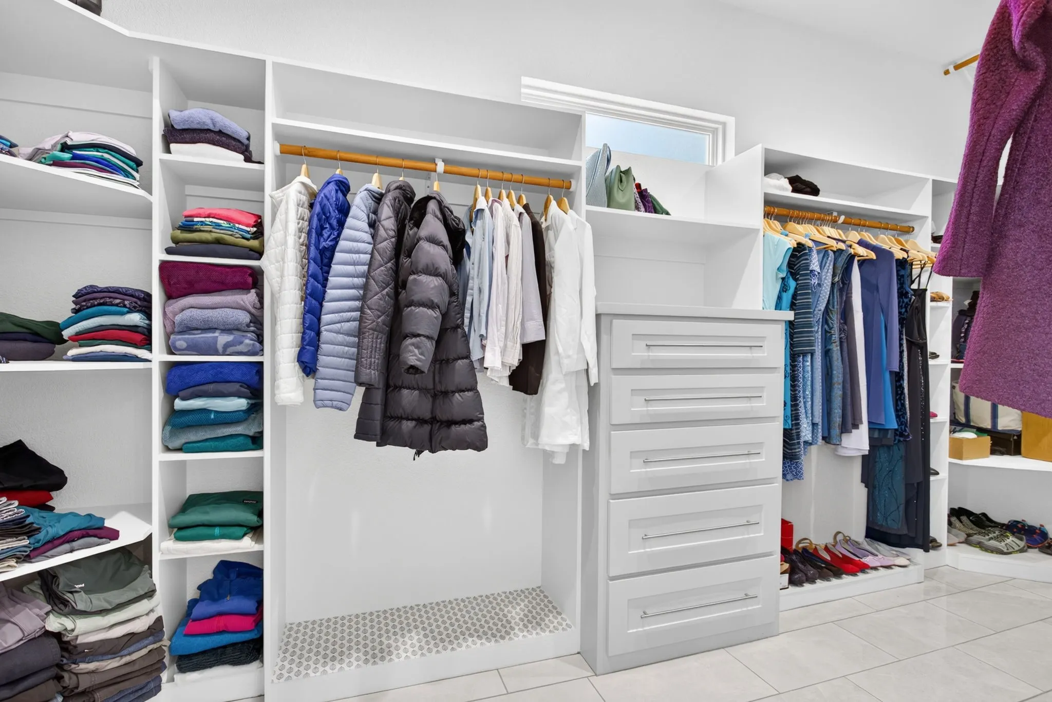 Expansive custom closet system featuring white shelving, multiple hanging rods, built-in drawers with modern hardware, and a window providing natural light