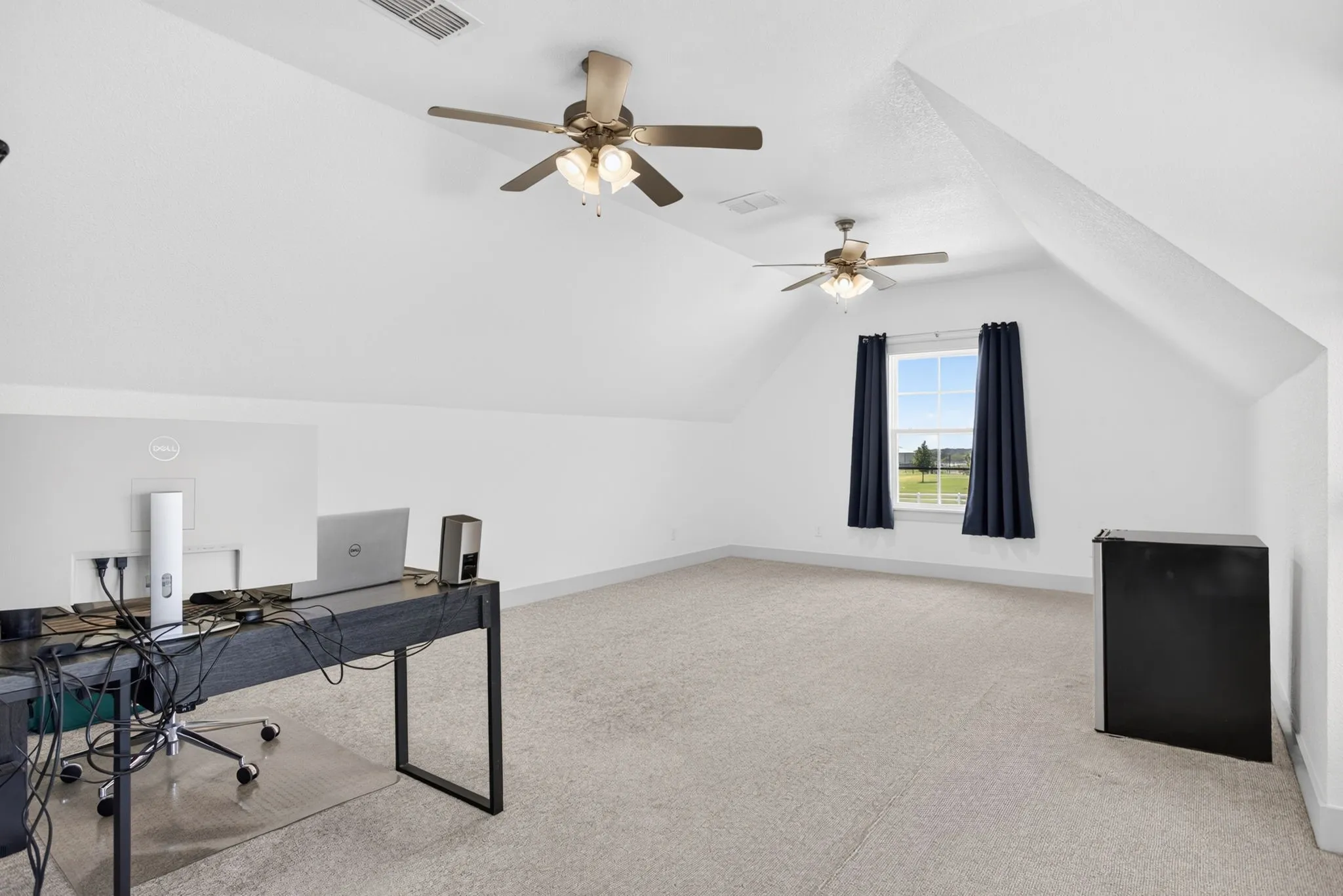 Spacious bonus room featuring vaulted ceilings, two ceiling fans with integrated lighting, window with dark curtains, and light-colored carpeting