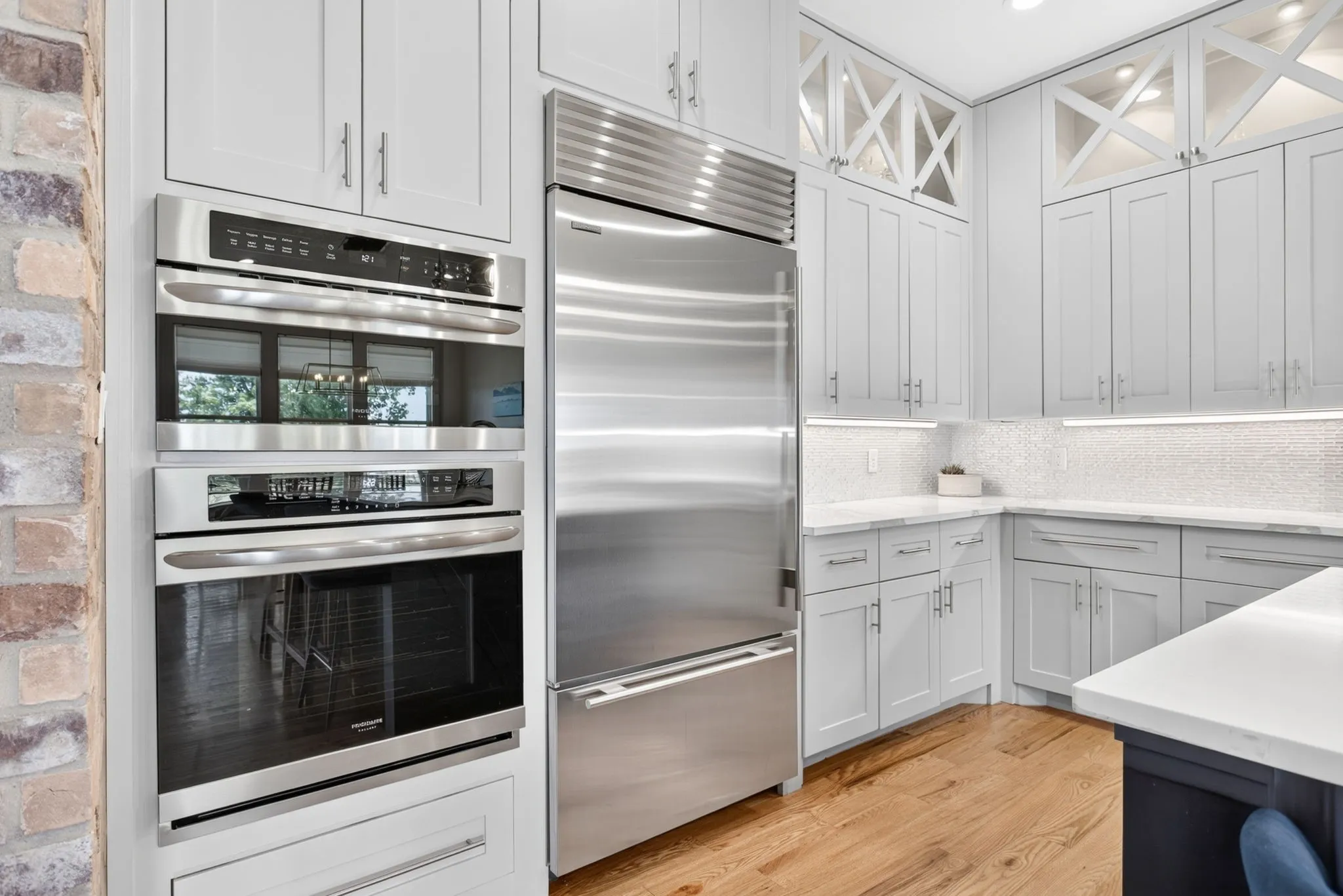 Gourmet kitchen featuring a built-in stainless steel refrigerator, stacked double ovens, light-toned cabinetry with glass accents, white countertops, and wood-finish flooring