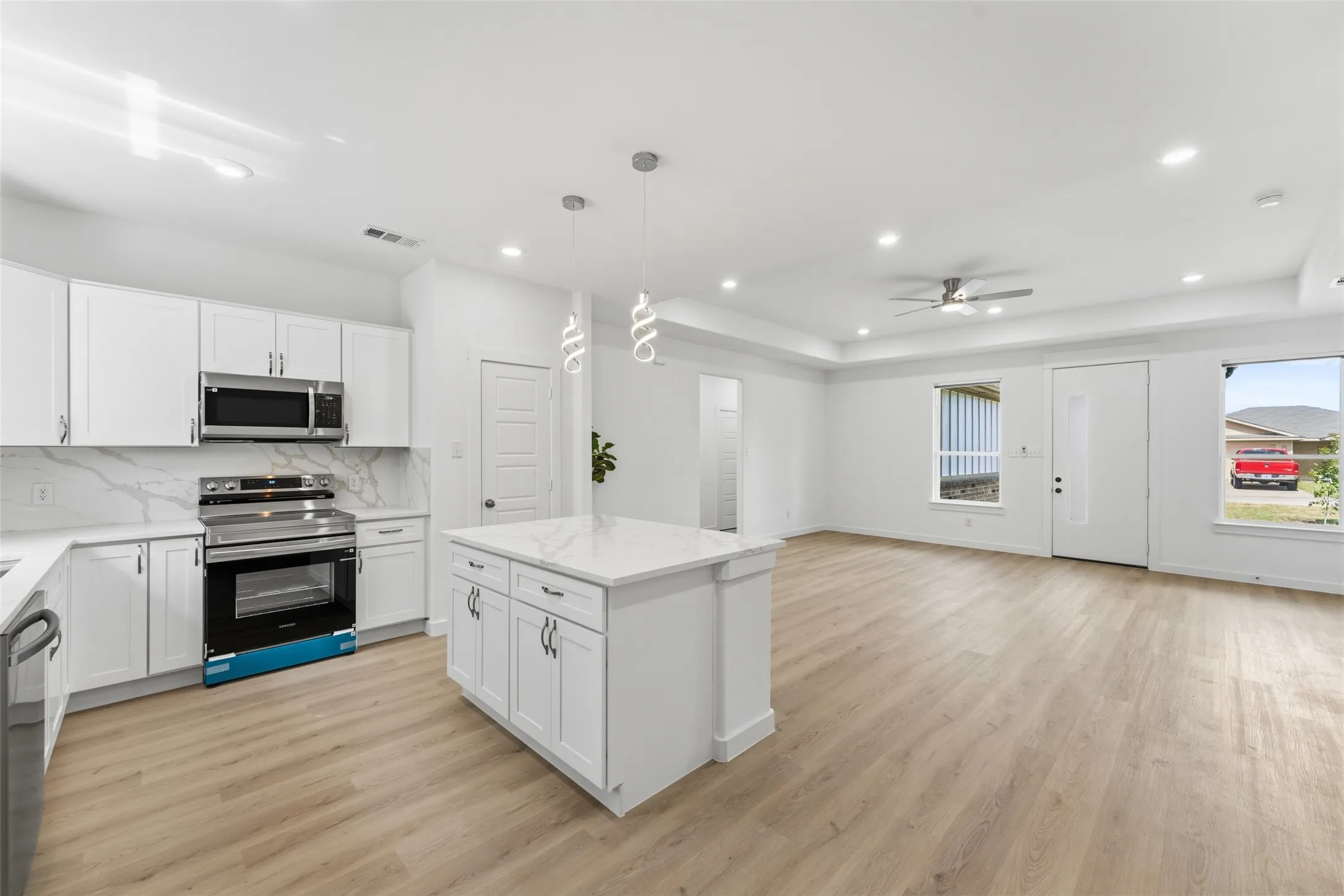 Modern kitchen and living area featuring a central island with pendant lighting, stainless steel appliances, white cabinetry with contemporary hardware, and wood-finish flooring