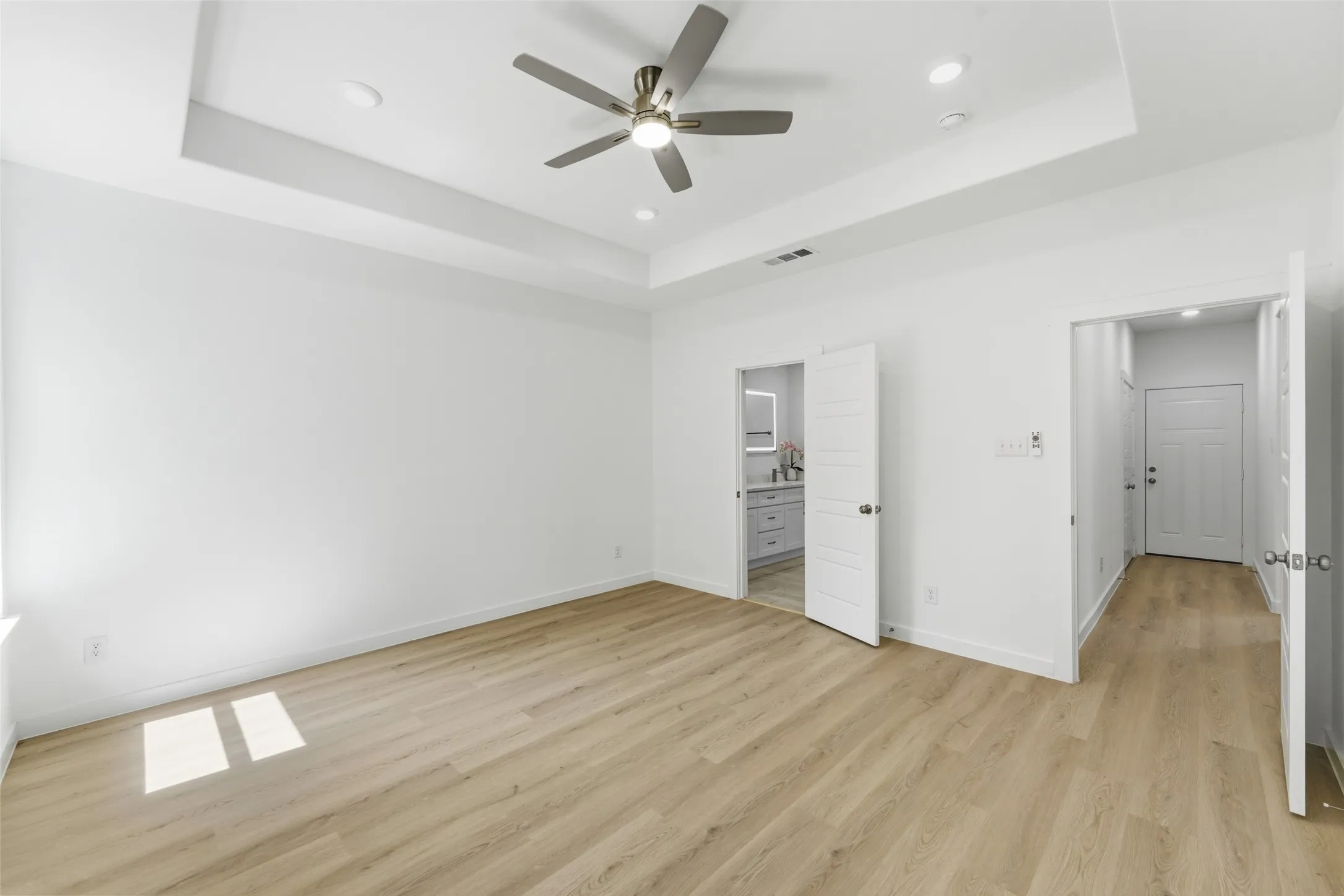 Spacious room featuring light wood-finish flooring, a tray ceiling with recessed lighting, and a modern ceiling fan