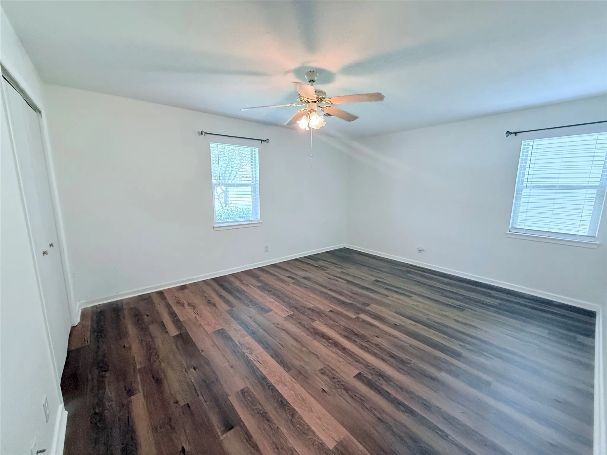 Spacious interior featuring wood-finish flooring, two windows with blinds, a ceiling fan with integrated lighting, and a bi-fold closet door