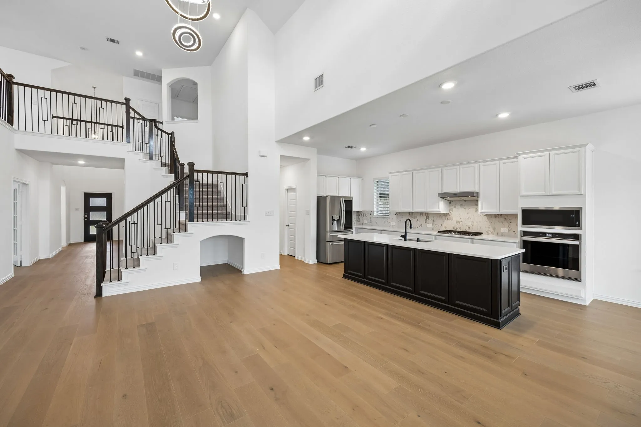 Expansive open-concept living area featuring a two-story ceiling, wood-finish flooring, a contemporary kitchen with a large island, stainless steel appliances, and a grand staircase with decorative balusters