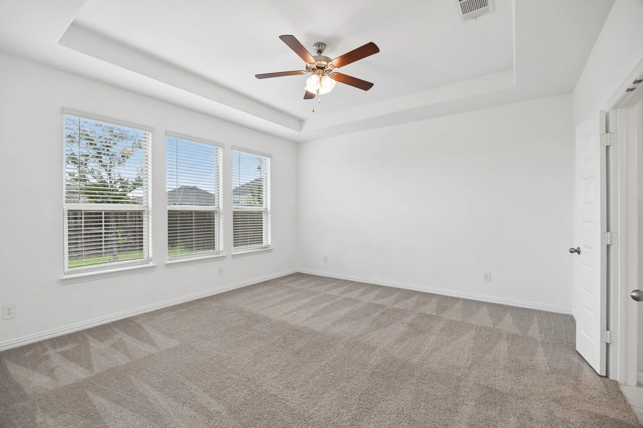 Bright room featuring three large windows with blinds, tray ceiling, ceiling fan with light fixture, and plush carpet flooring