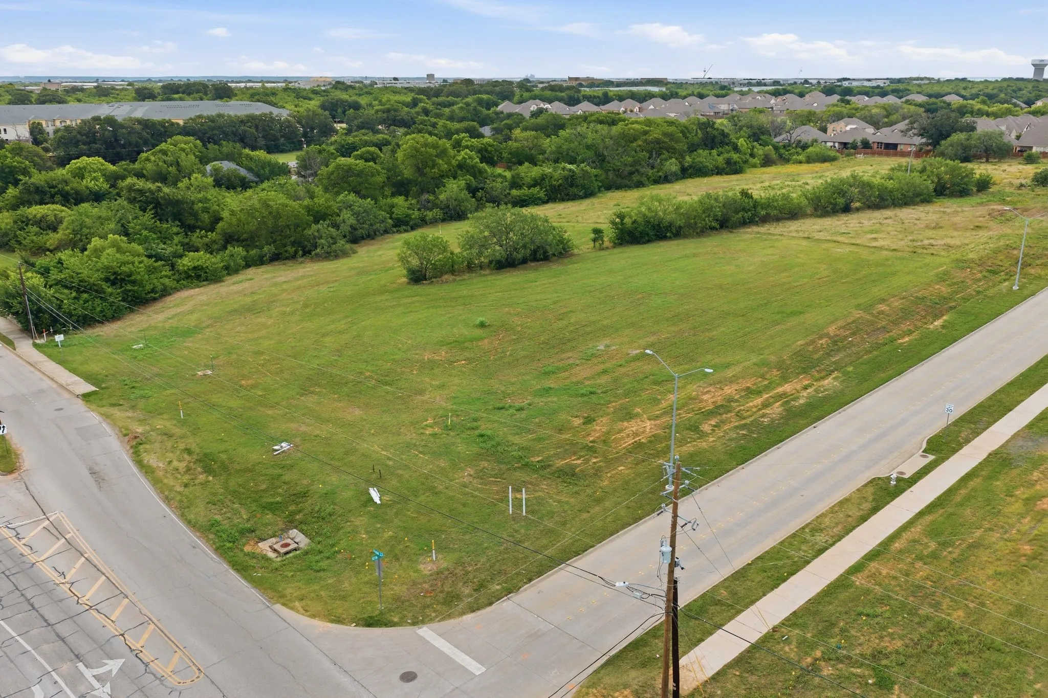 Expansive corner lot featuring a large grassy area, mature trees bordering the property, and direct frontage on two paved roads with sidewalks